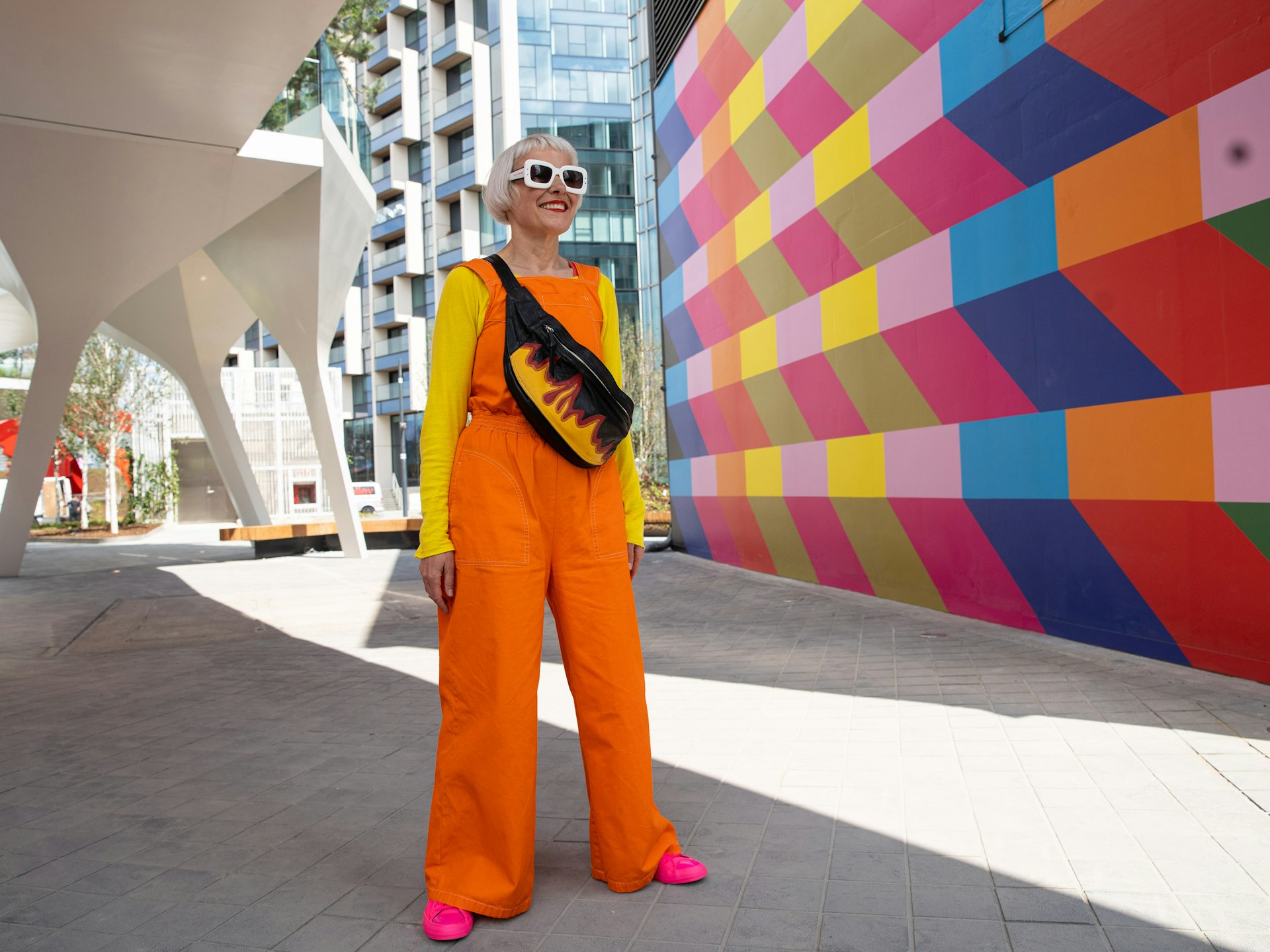 Woman dressed in bright jumpsuit stood  under footbridge by bright multicoloured wall