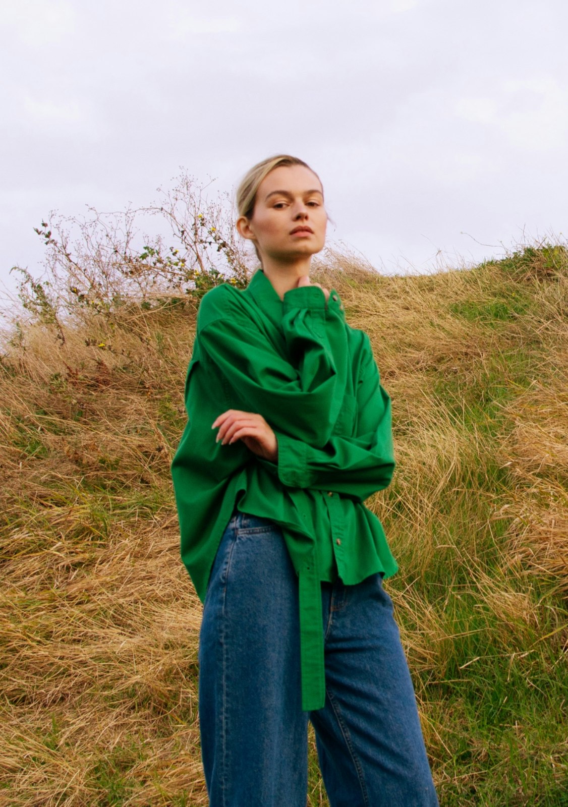 A lady stands in a field wearing a green blouse and blue jeans
