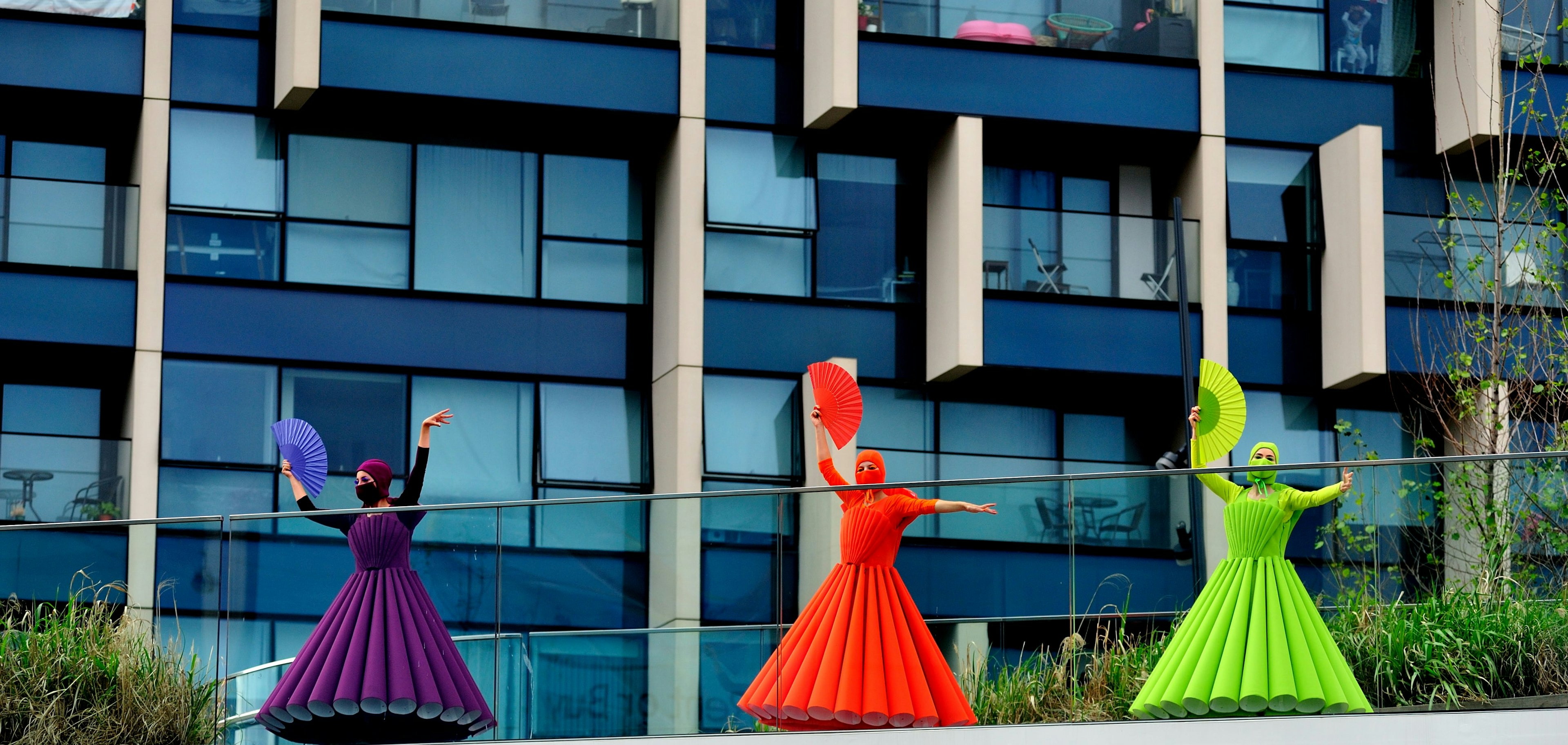 3 people dressed in single bright coloured dresses performing a dance with a Greenwich Peninsula building in the background