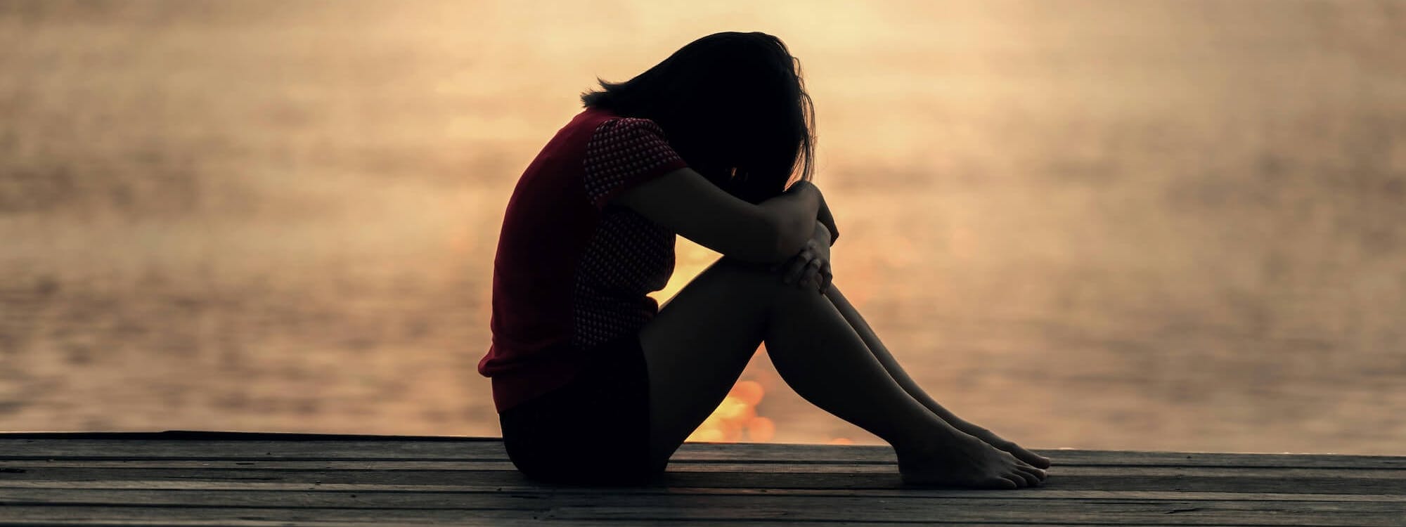 Frau sitzt betroffen auf einem Steg am See