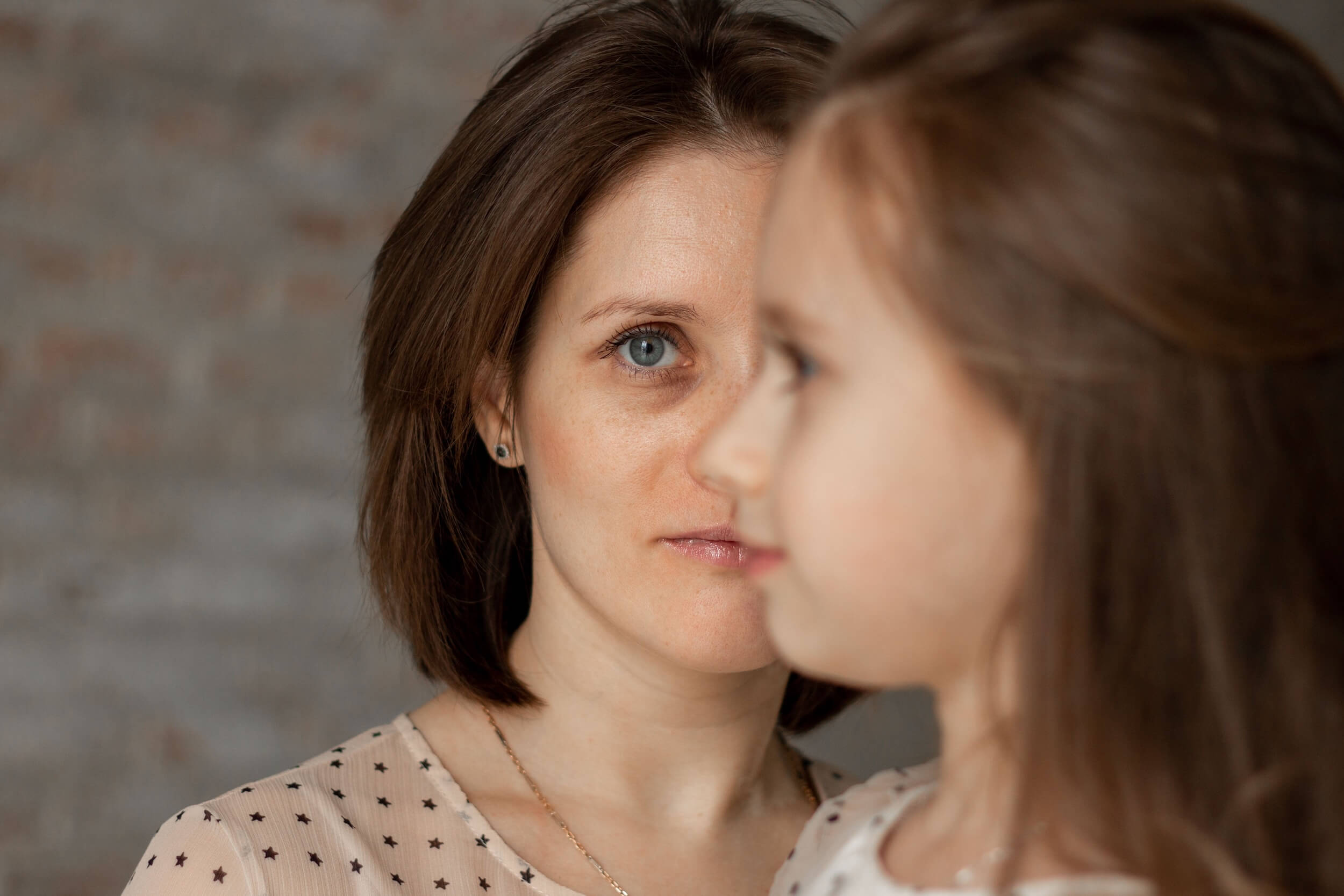Mutter im Halbprofil, den Blick nachdenklich auf ihre kleine Tochter gerichtet, die unscharf im Vordergrund steht