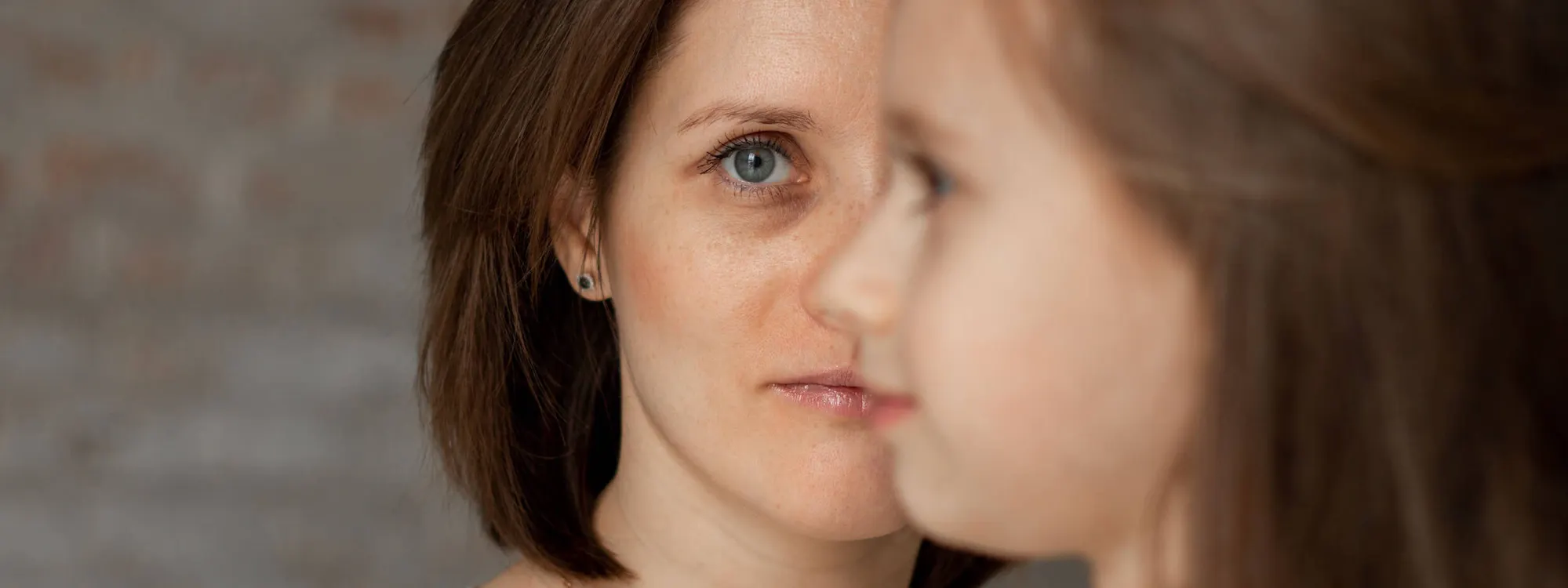 Mutter im Halbprofil, den Blick nachdenklich auf ihre kleine Tochter gerichtet, die unscharf im Vordergrund steht