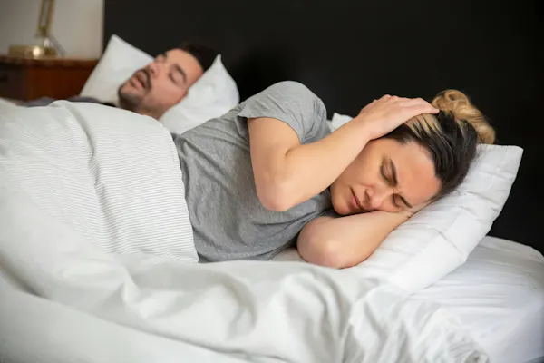 Frau liegt im Bett und hält sich die Ohren vor Schmerzen