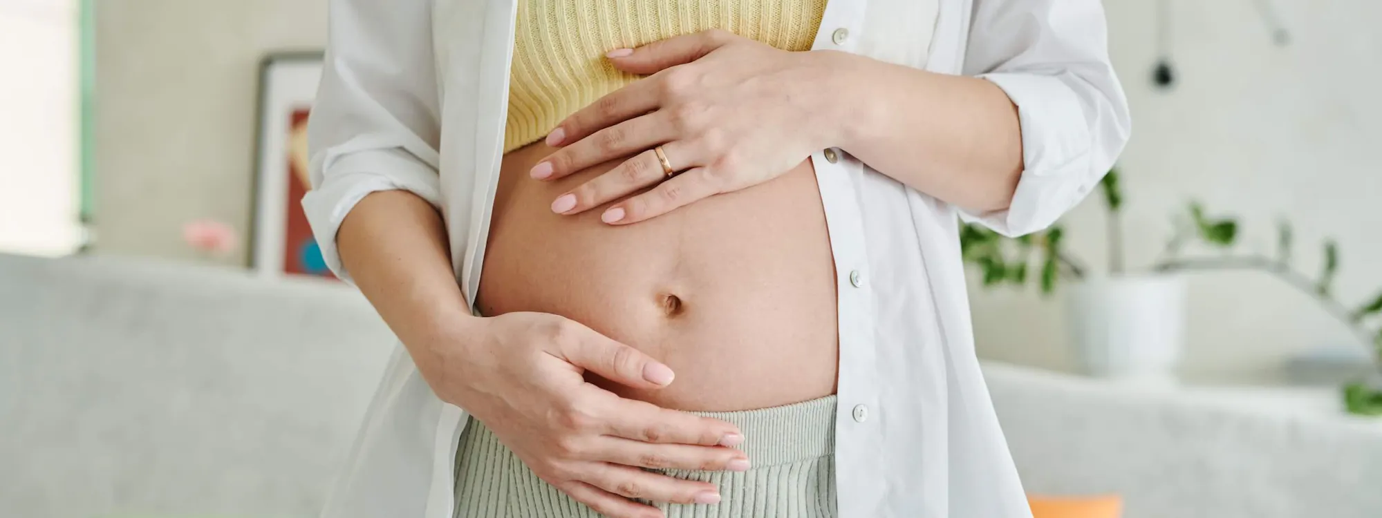 Schwangere Frau steht im Wohnzimmer und hält sich die Hände an den Bauch