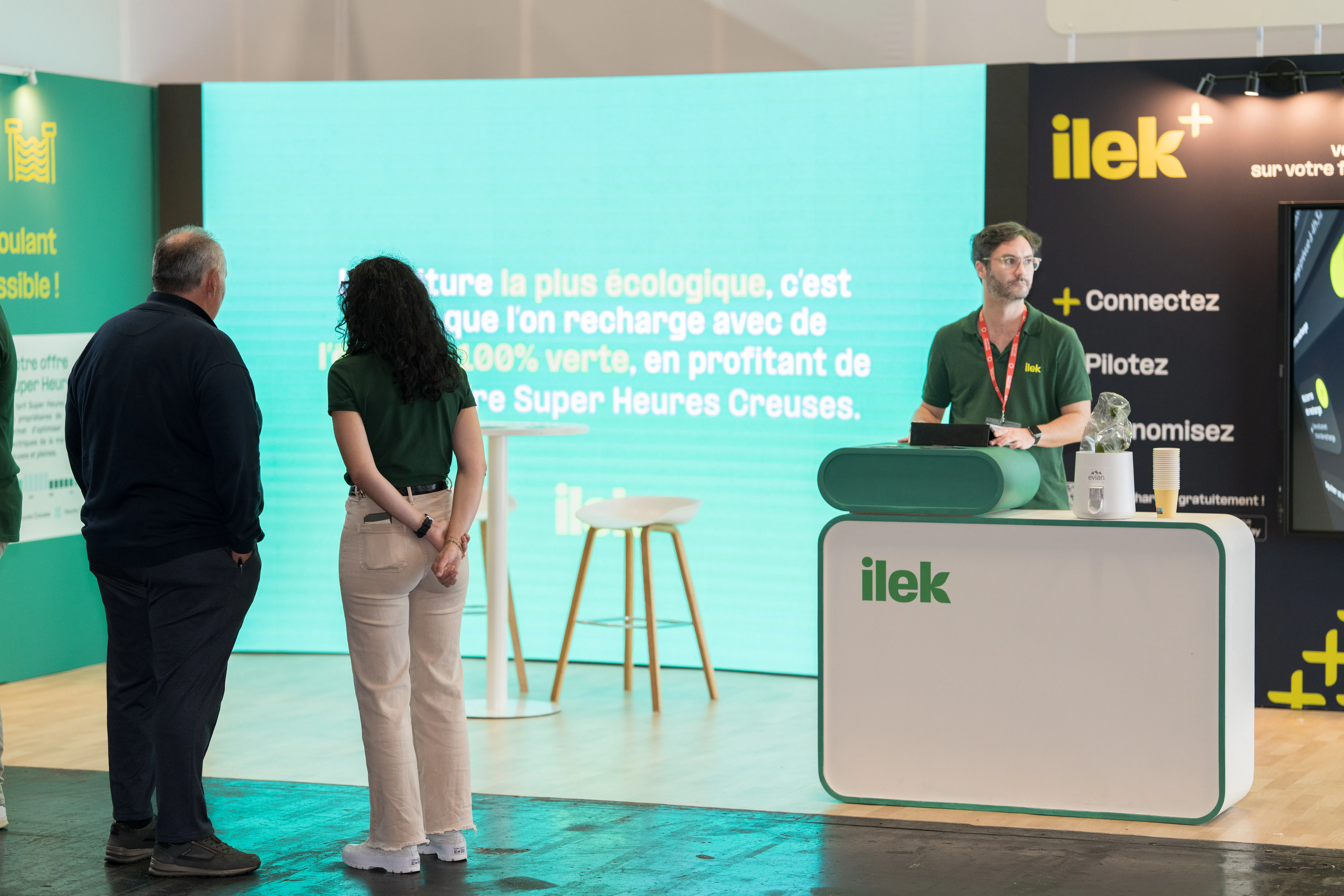 Hôtesse Ilek échangeant avec un visiteur sur l’offre de recharge de véhicules électriques et l’énergie verte, stand conçu par notre agence événementielle.