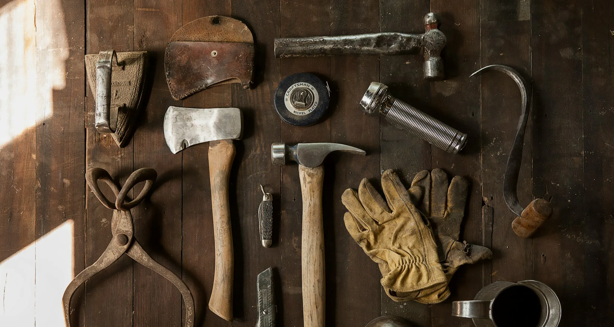 Hammer, Beil, Maßband und Arbeitshandschuhe mit weiteren Werkzeugen auf dunklen Holzplanken.