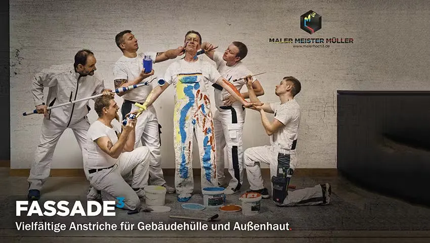 Gruppe in weißer Arbeitskleidung gestaltet eine bunte Figur, darunter der Schriftzug „Fassade“.