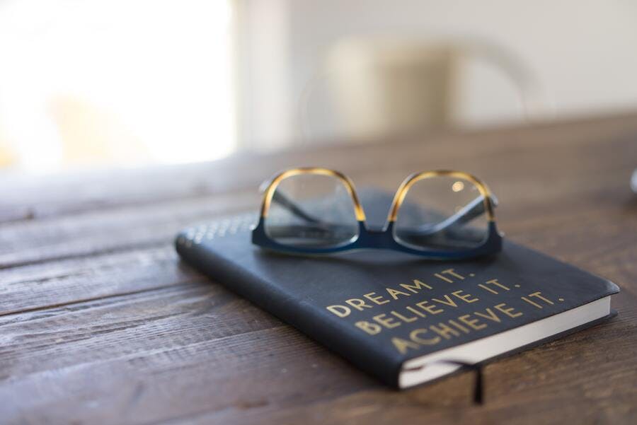 A book and a pair of glasses on top of a wooden table. 