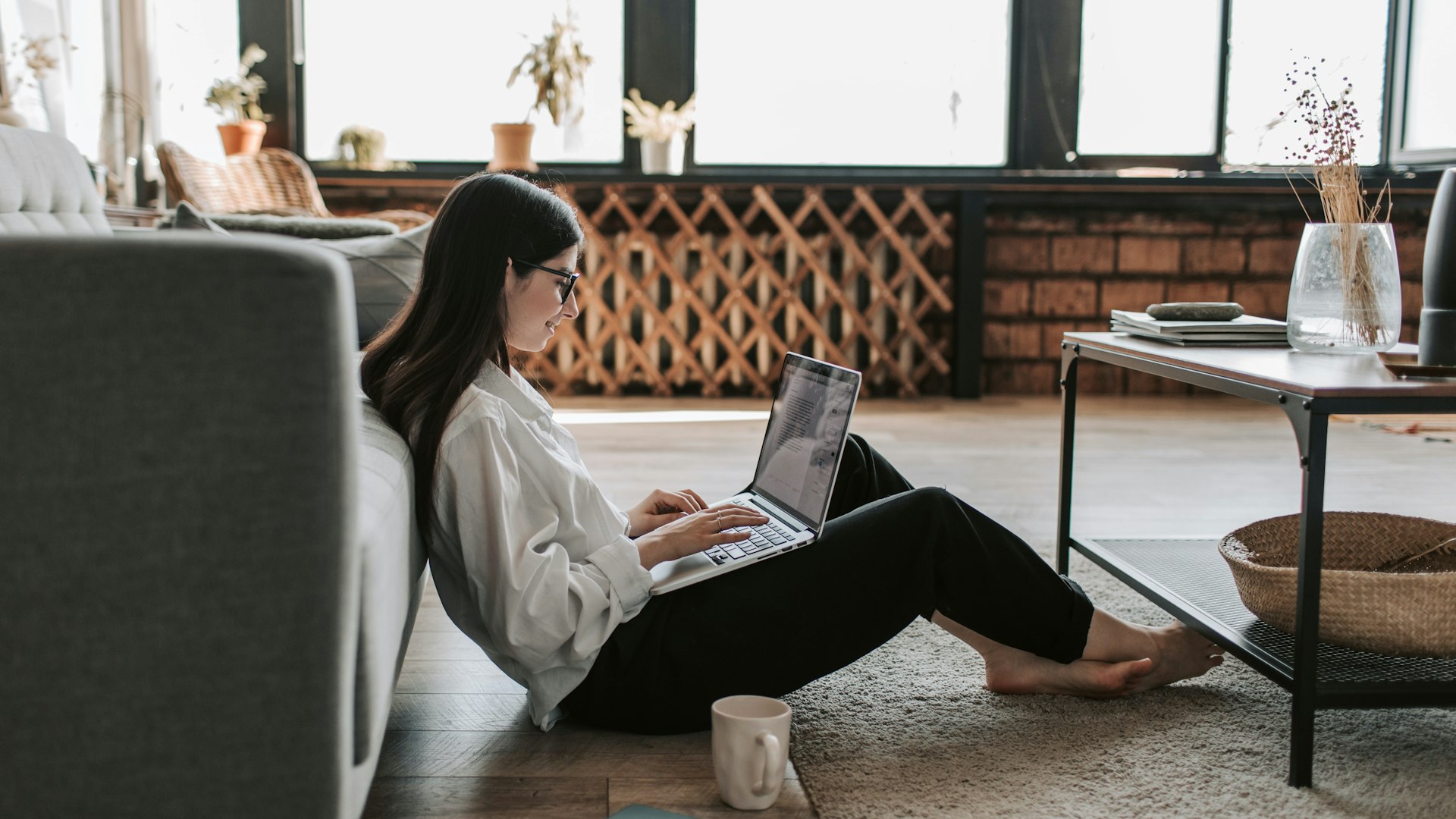A woman sitting on the floor, in between the sofa and the living room table, with a laptop in her lap and a coffee cup next to her.