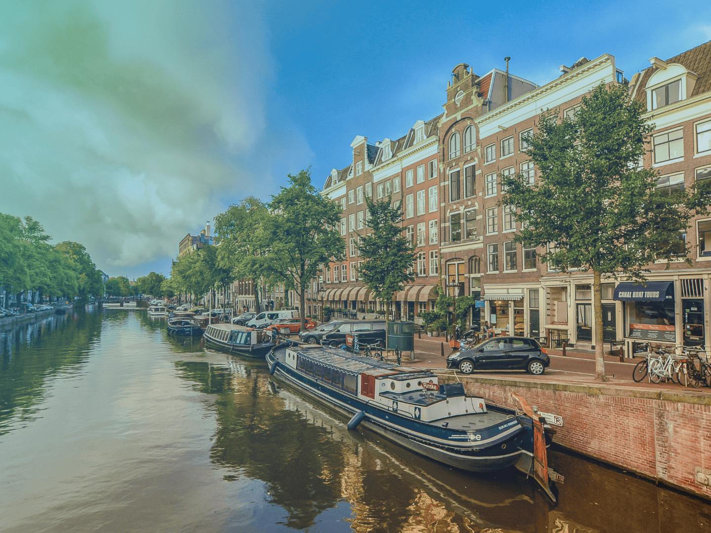 Hollanda'da kanala karşı dizilmiş binalar. Tekneler park edilmiş. İnsanlar kanal kenarında bisiklet sürüyor. 