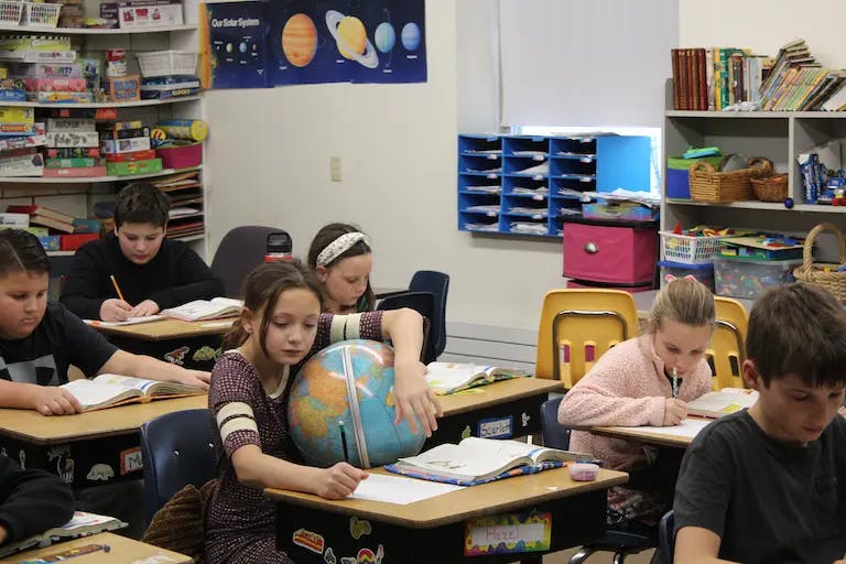 Students at desks writing, and the student in the middle is holding a globe while writing