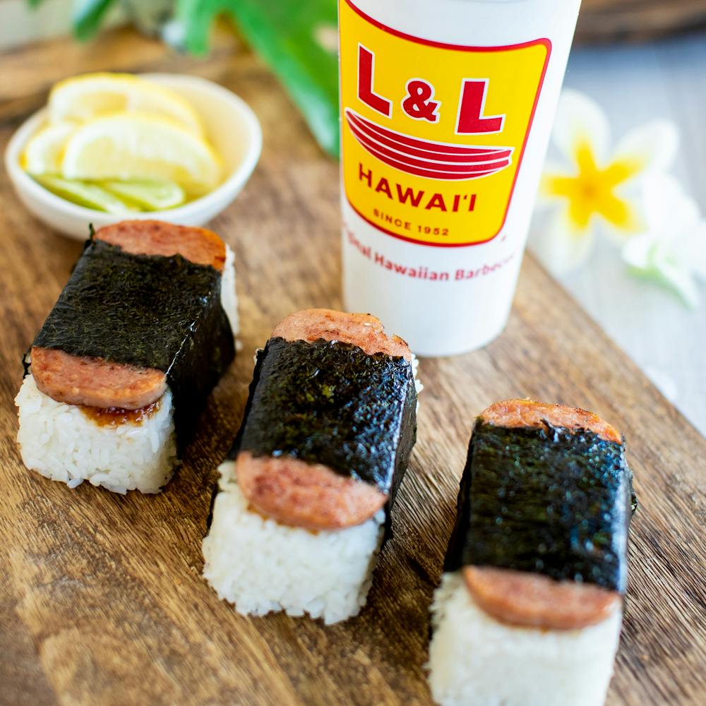 An image of three SPAM® musubi and an L & L cup with lemon wedges and a plumeria flower in the background.
