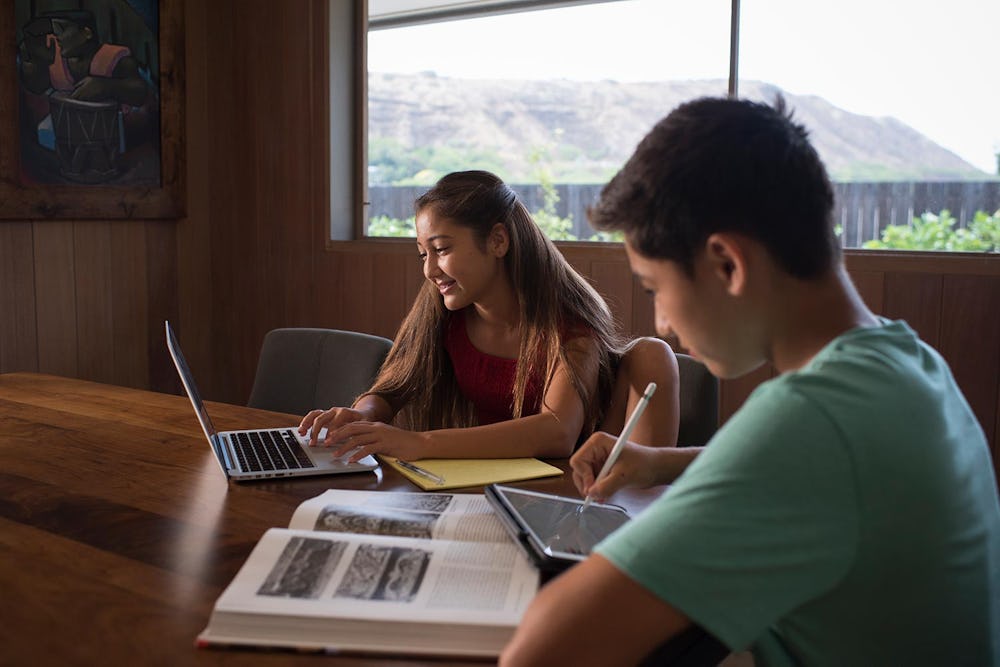 Boy and girl studying in living room with a laptop, tablet, and book.