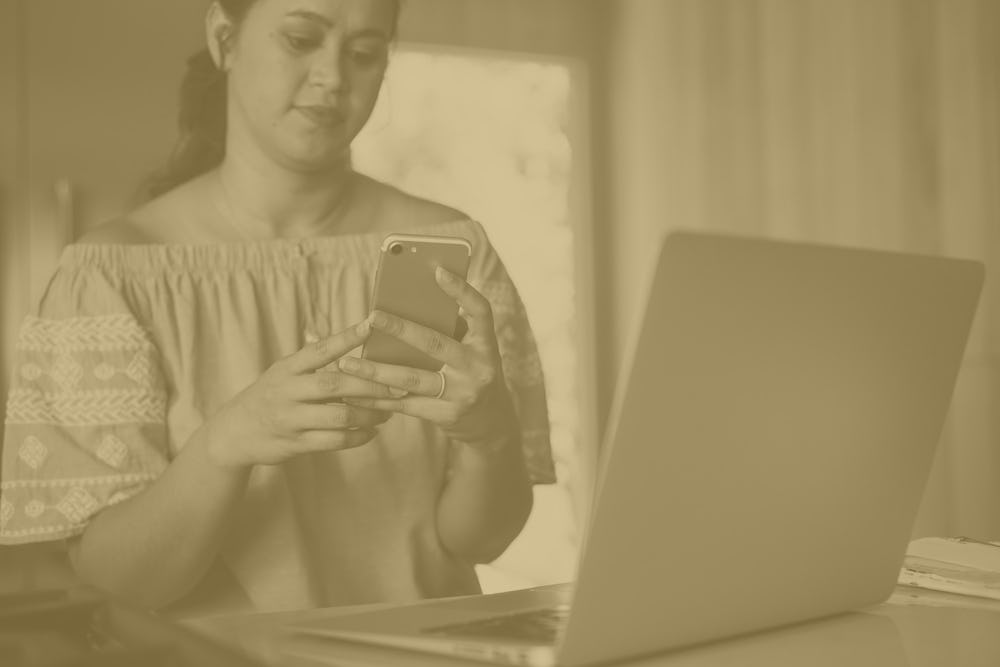 woman looking at cell phone with laptop in front of her