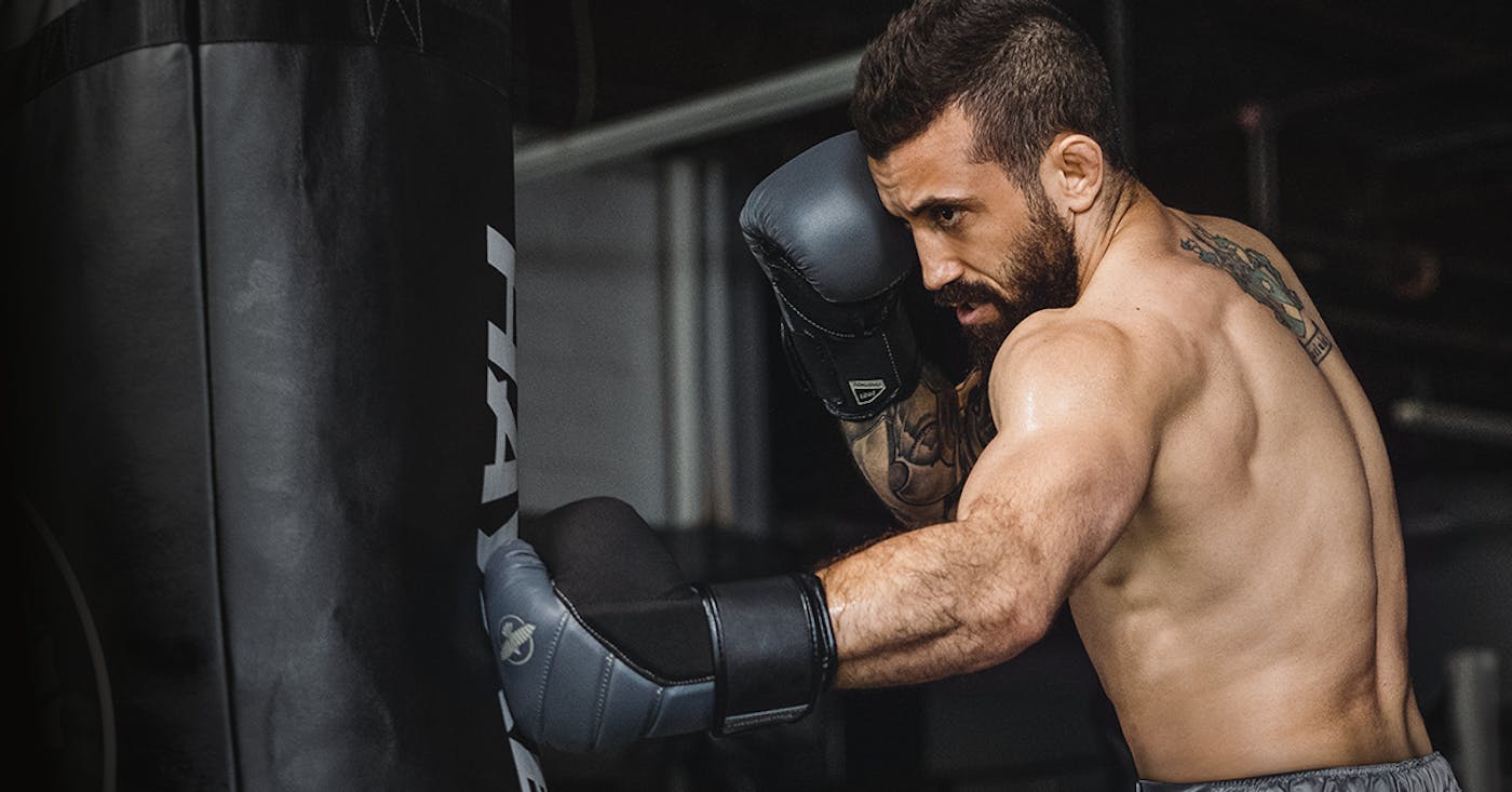 A boxer punching a heavy bag in charcoal colored boxing gloves to reveal his shoulder and back muscles.