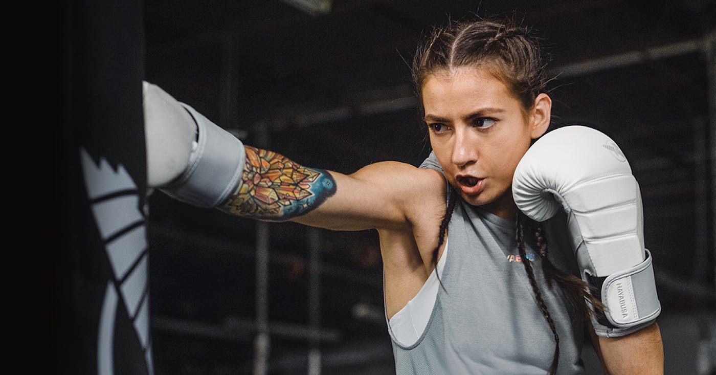 A woman in a boxing gym punching a heavy bag with white/grey Hayabusa T3 Boxing Gloves.