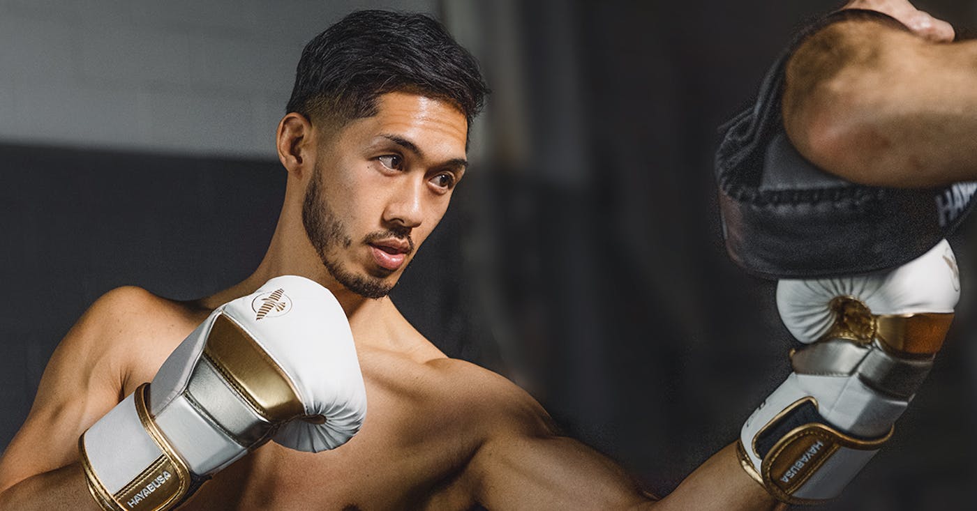 A man in a boxing gym in fighting stance punching a thai style pad with white/gold Hayabusa T3 Boxing Gloves.