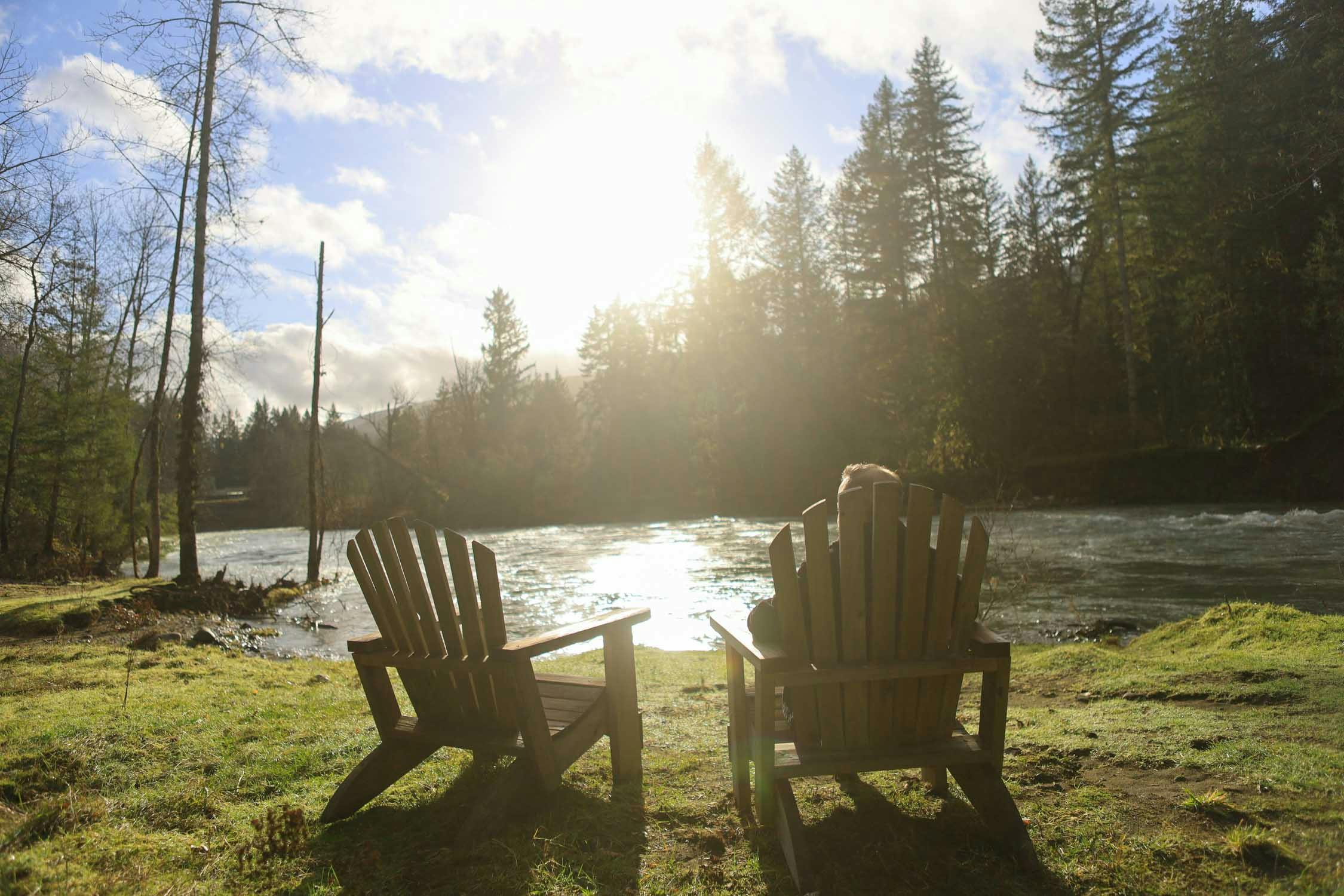 A photo of two wooden chairs facing the river with the sun shining and sparkling reflections on the water