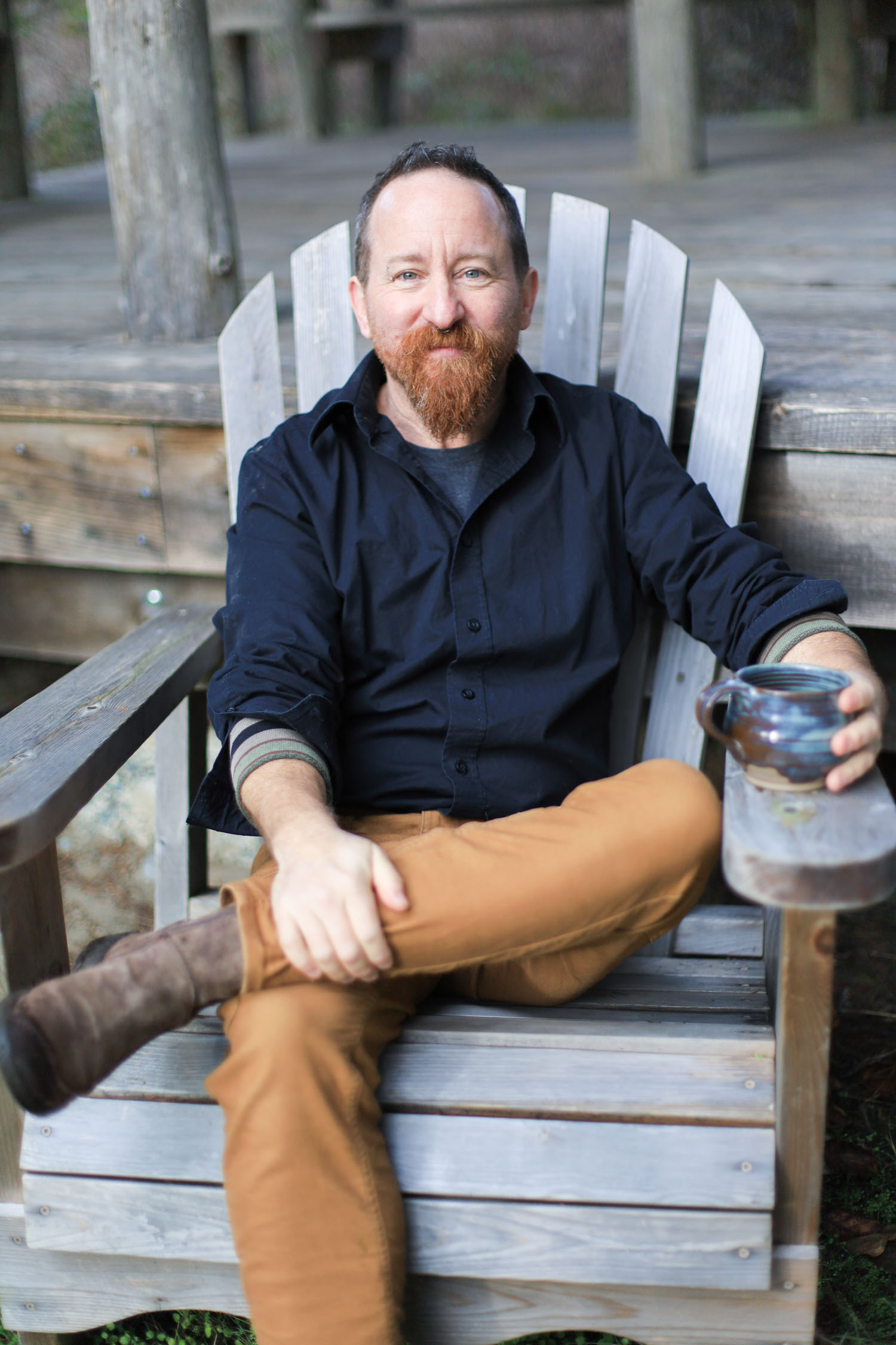A photo of dean sitting cross legged in a chair outdoors, holding a tea mug with a welcoming expression