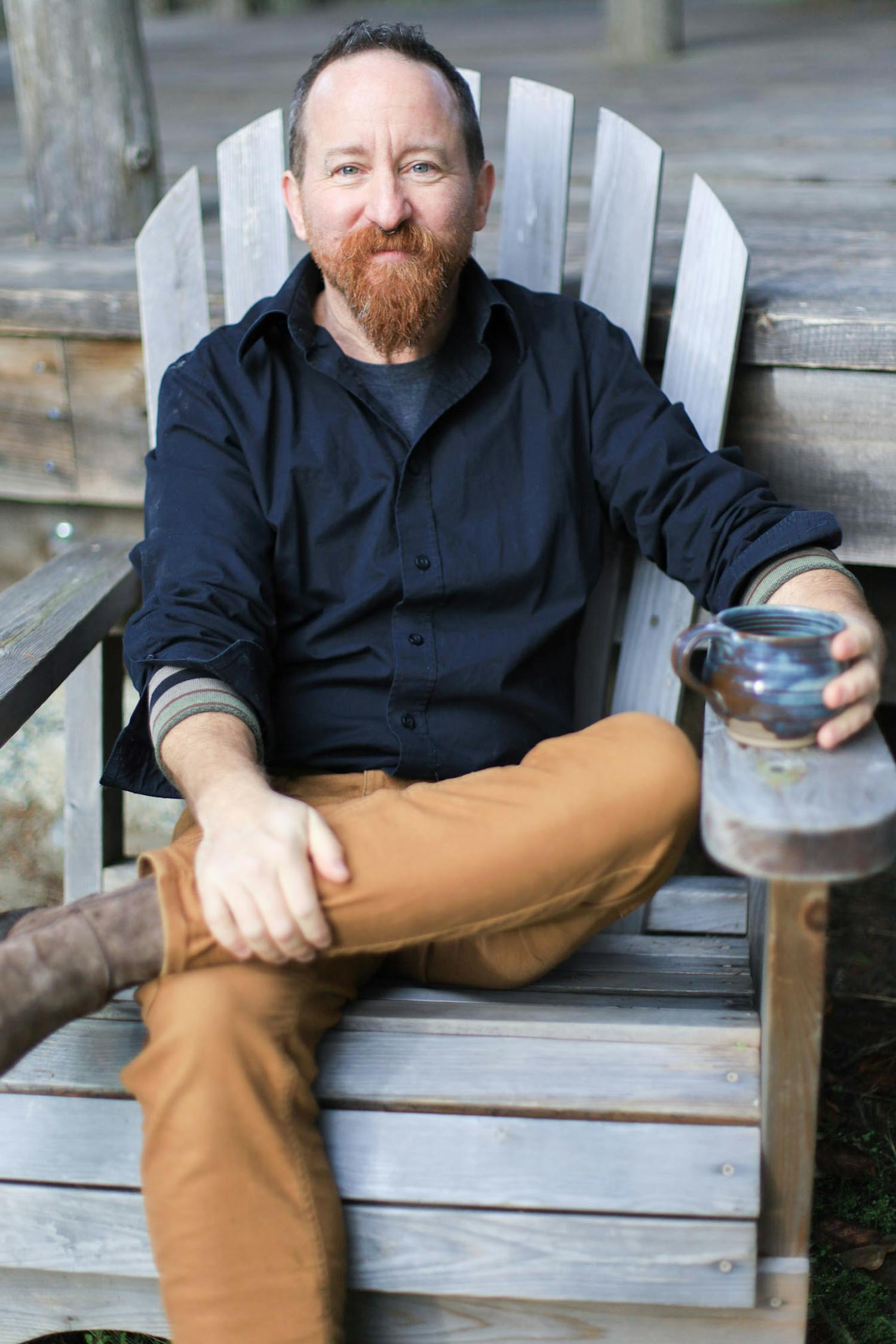 A photo of dean sitting cross legged in a chair outdoors, holding a tea mug with a welcoming expression