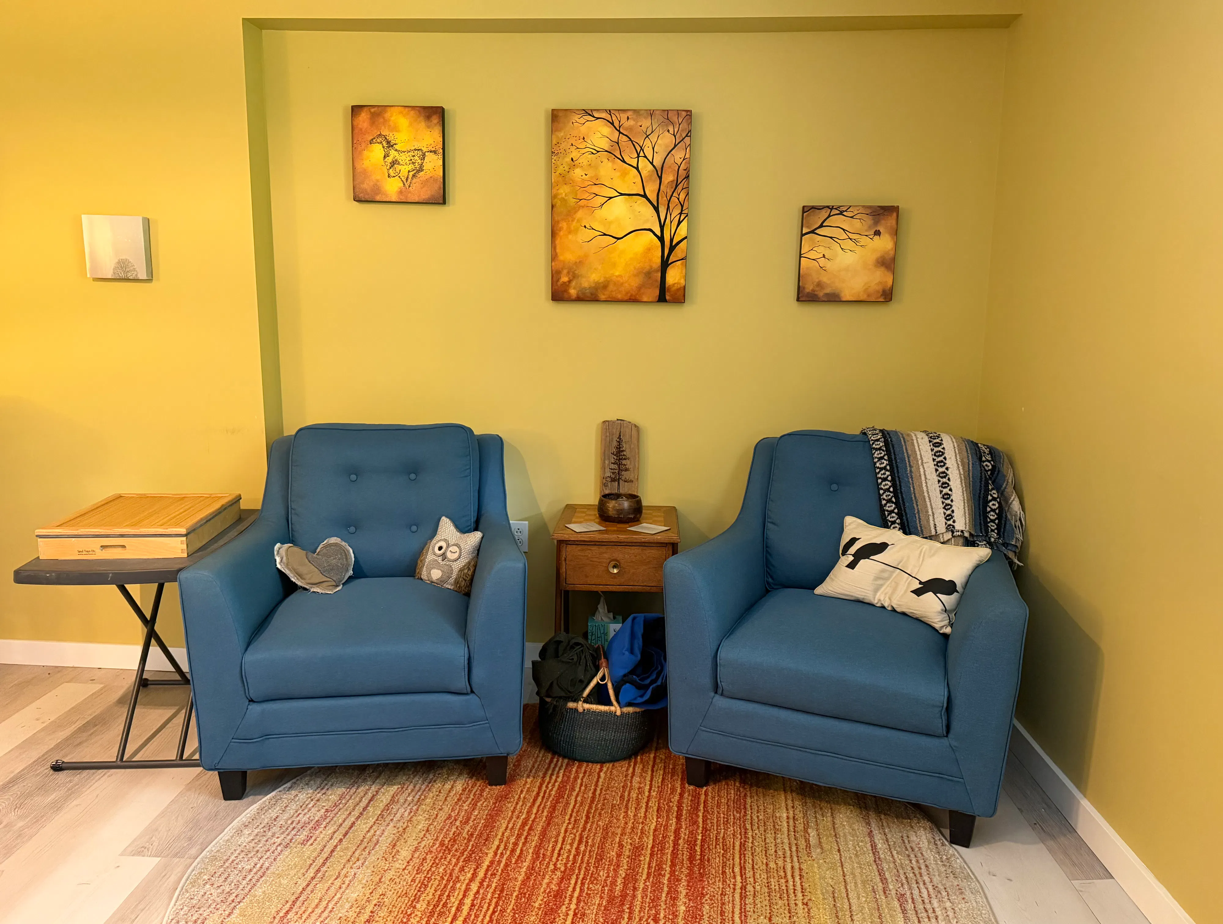 A photo of an interior room with two blue chairs, yellow walls, a rug, and a paining of a tree.