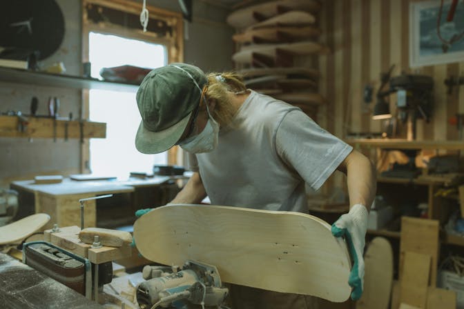 Handwerker mit Kappe und Mundschutz schleift Holz für ein Skateboard in einer Werkstatt ab