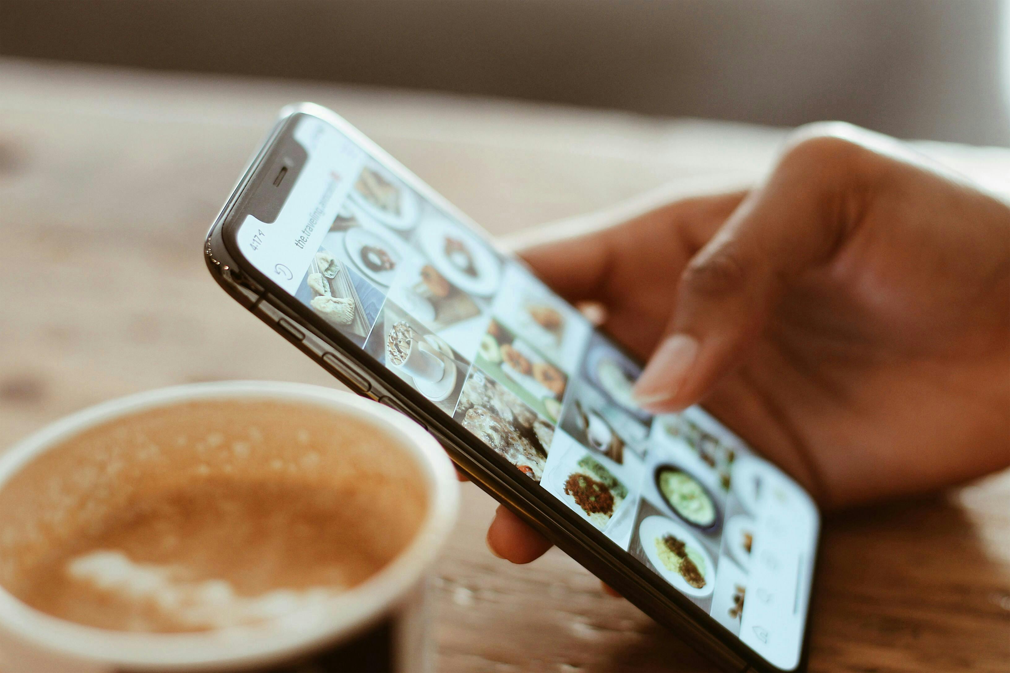 A Right Hand is resting on a table. The hand is holding a phone that shows the Instagram Explore Page. In the foreground, out of focus, is a Cup of Coffee in a brown to-go cup.