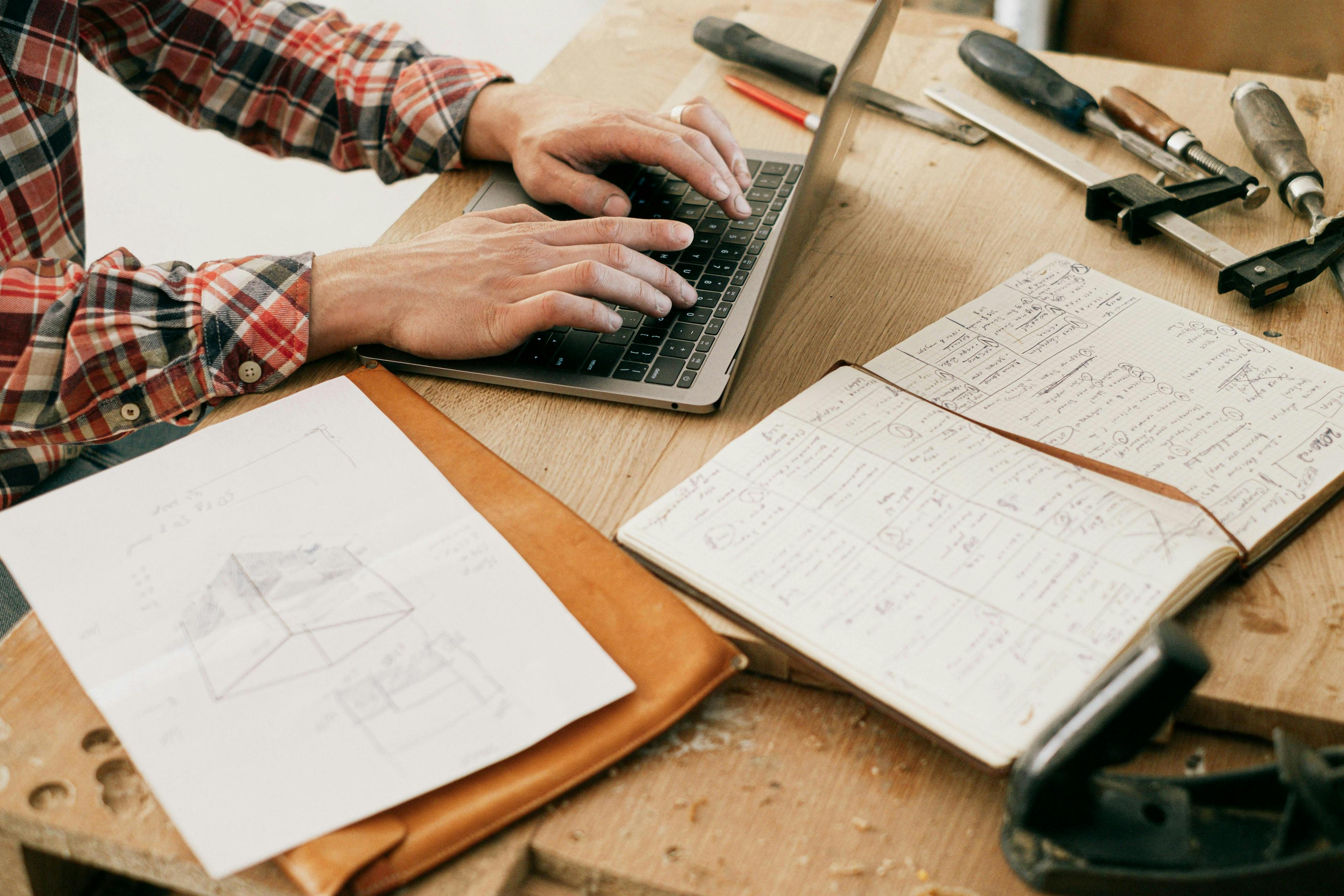 man typing on a laptop ontop a table with various hand written notes and sketches surrounding him, on his left side are a few tools