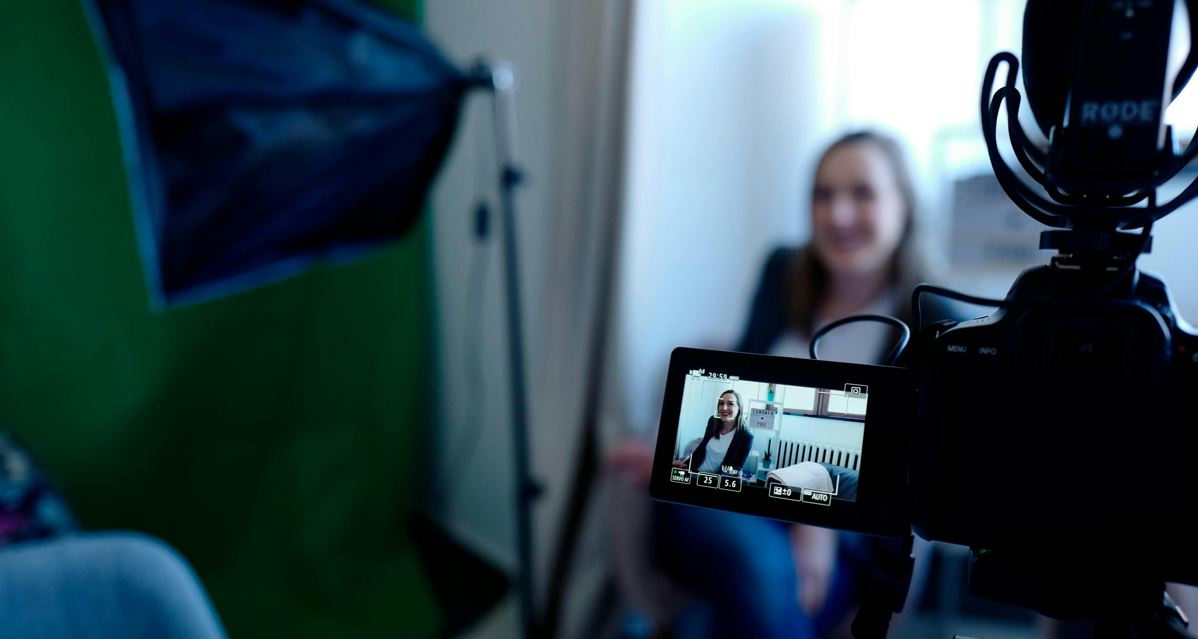 Woman sitting in a filming studio out of focus. The focus is on a video camera showing the woman in focus on the view finder