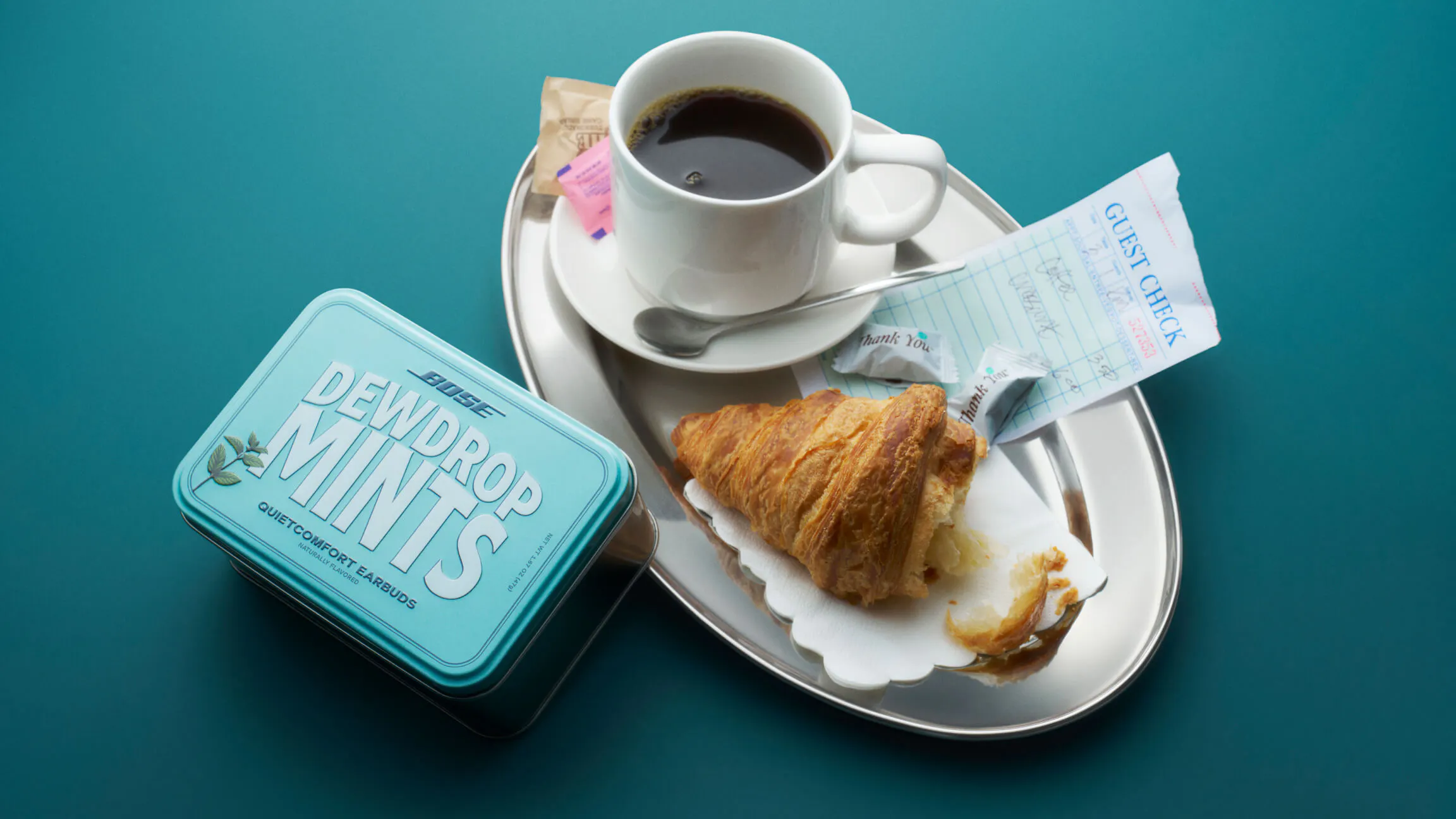 The Bose QuietComfort Earbuds "Dewdrop Mints" seeding tin on a teal surface beside a diner tray holding a black coffee, a half-eaten croissant, sugar packets, and a guest check.