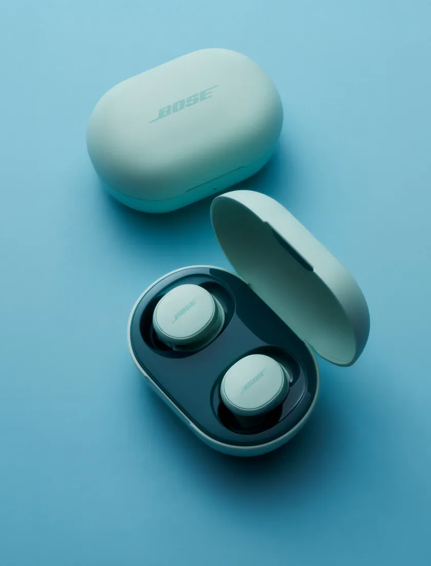 Bose QuietComfort Earbuds in Dewdrop Mint colorway. The open charging case sits in the foreground with both earbuds seated inside; the closed case lid rests behind it on a light blue surface.