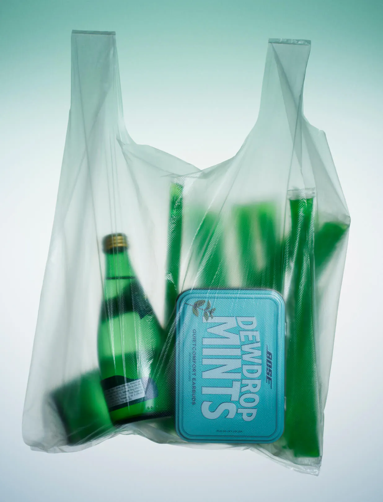 The Bose Dewdrop Mints tin inside a translucent plastic grocery bag alongside a green glass bottle and green celery stalks, shot against a pale mint background.