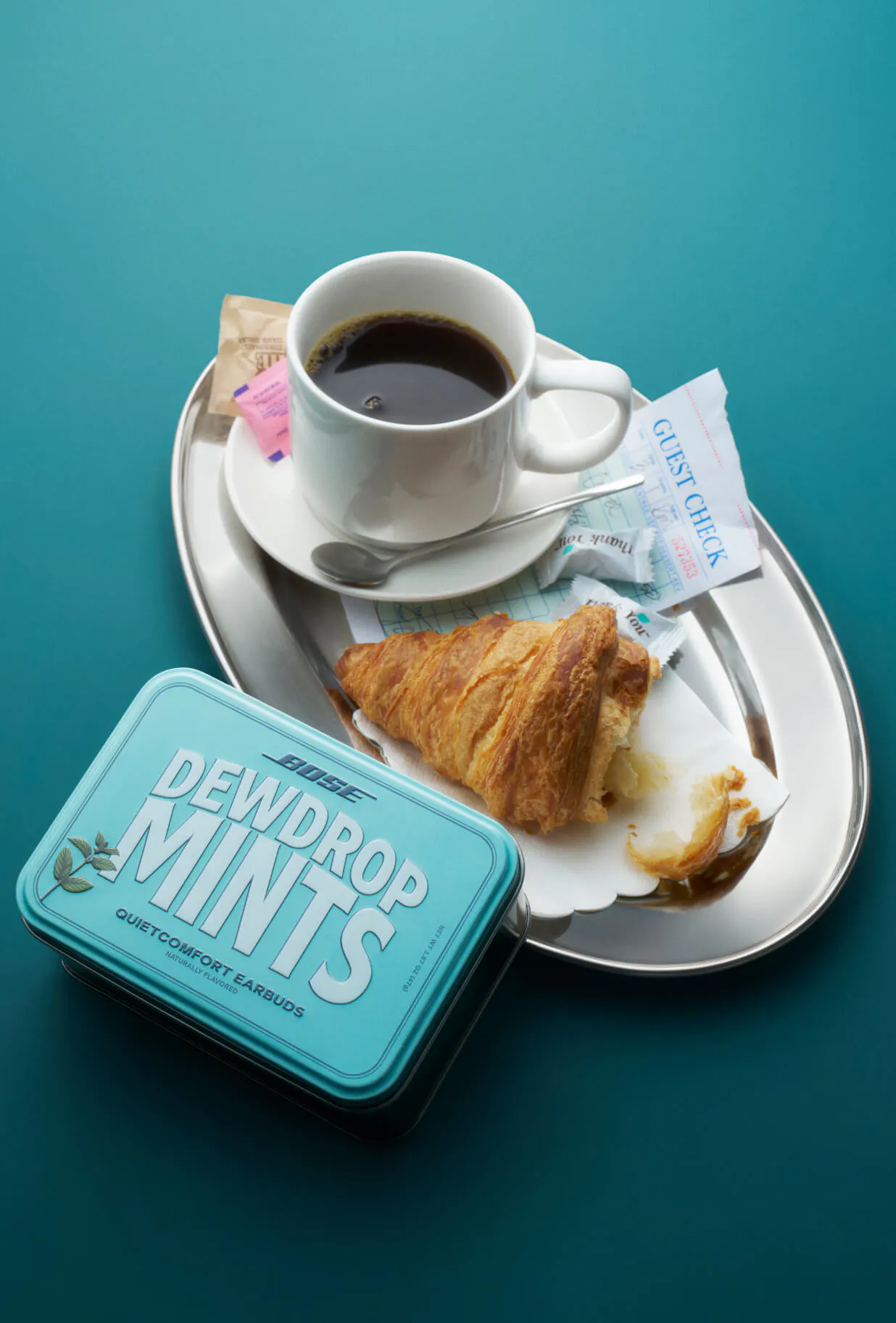 The Bose QuietComfort Earbuds "Dewdrop Mints" seeding tin on a teal surface beside a diner tray holding a black coffee, a half-eaten croissant, sugar packets, and a guest check.