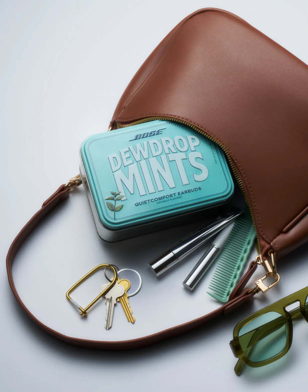 The Bose Dewdrop Mints tin half-tucked into an open tan leather bag, surrounded by spilled contents: a set of keys, two lip products, a mint-green comb, and olive-tinted sunglasses.