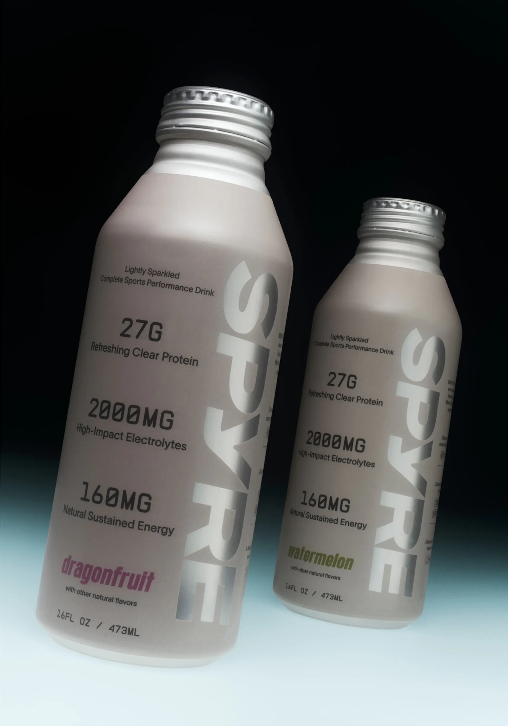 Two SPYRE performance drink bottles side by side—dragonfruit and watermelon flavors—in matte grey aluminum with silver caps, against a dark gradient background