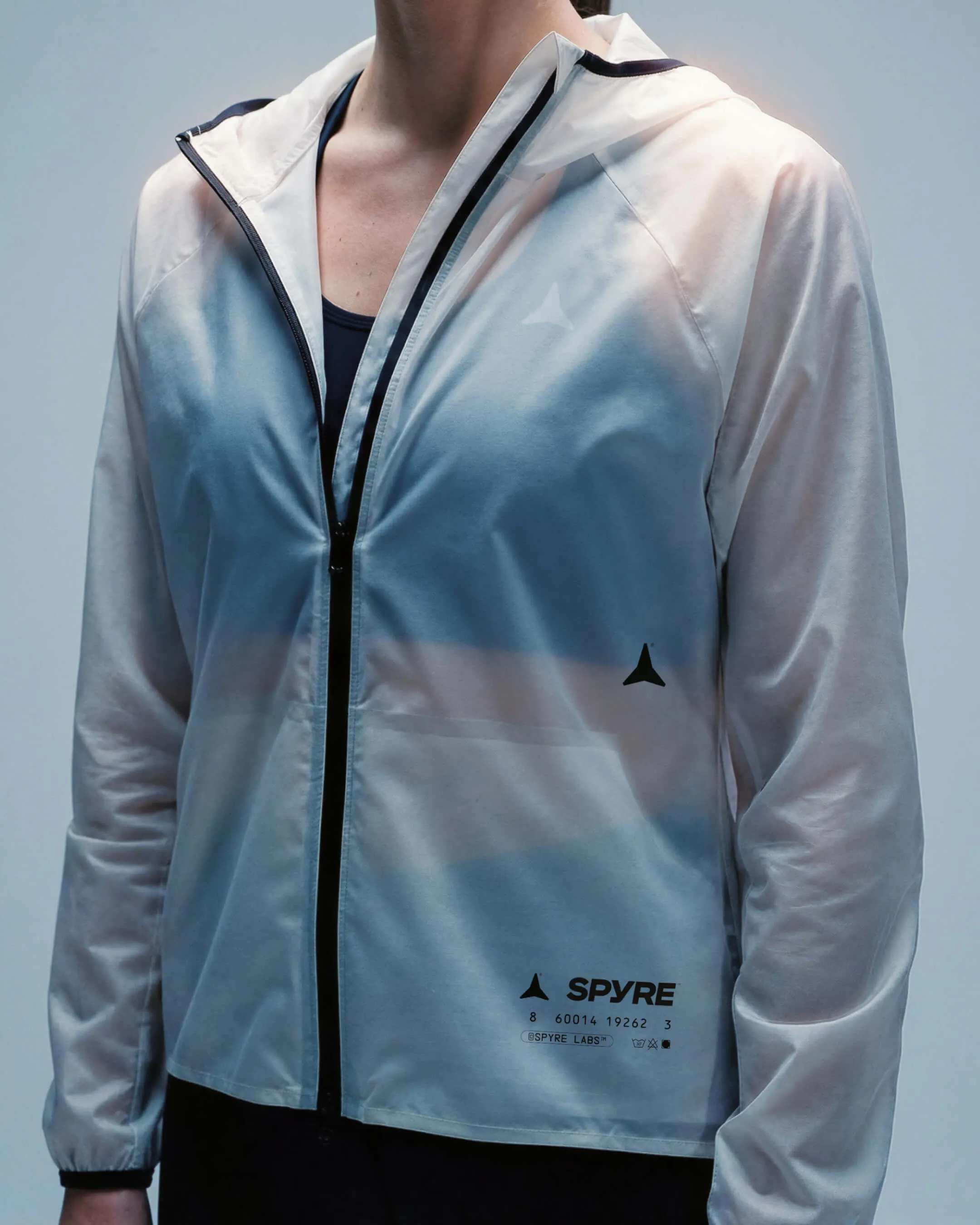 Model wearing a translucent white hooded windbreaker with SPYRE logo and barcode printed at the hem, worn open over a dark athletic top against a pale blue background