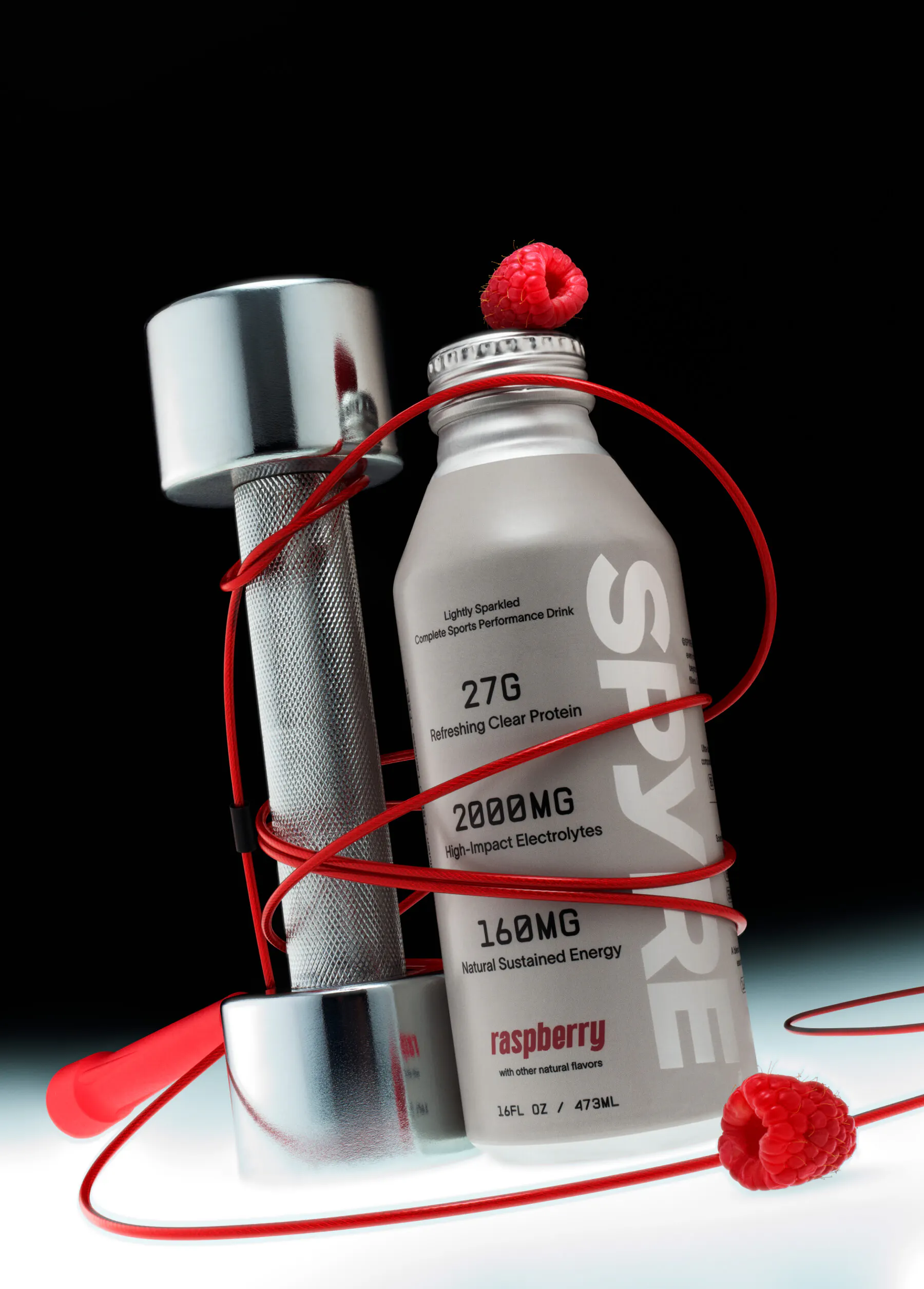 SPYRE raspberry performance drink bottle standing upright alongside a chrome dumbbell, wrapped in a looping red cord with fresh raspberries, against a dark gradient background