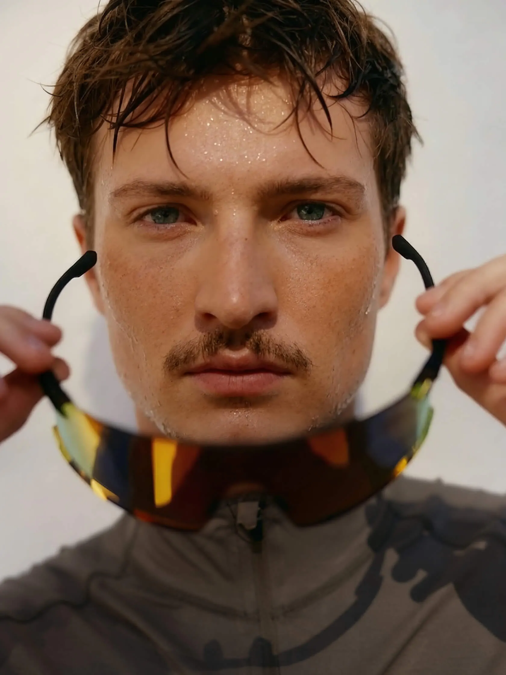 Close-up portrait of a sweaty male athlete holding mirrored sport sunglasses below his chin, wearing a patterned athletic top