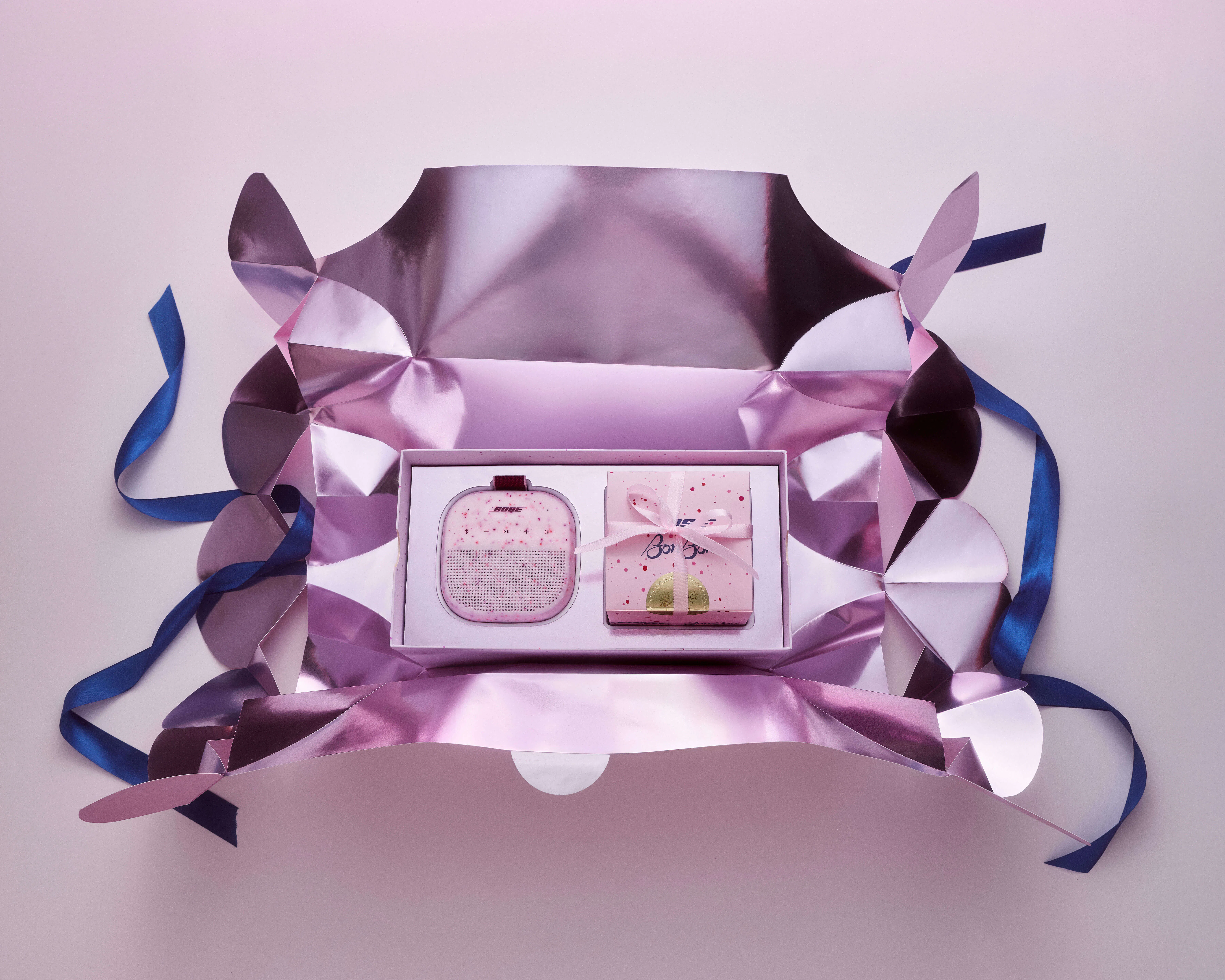 Overhead view of the fully opened Bose × BonBon candy wrapper mailer, its metallic pink interior and fanned paper ends splayed flat, revealing the inner gift box with the Soundlink Micro and BonBon candy box side by side.