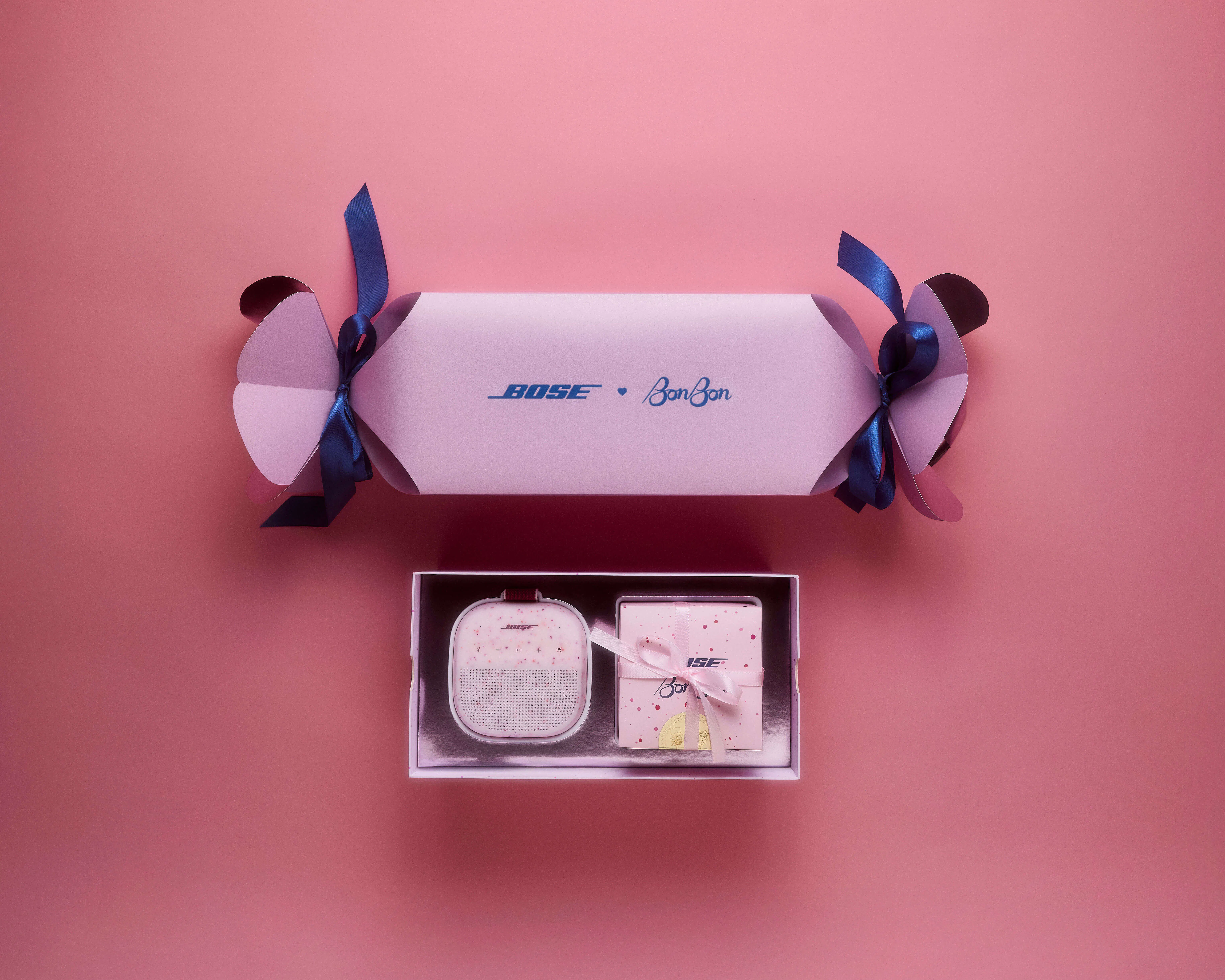 Flat lay of the Bose × BonBon candy wrapper mailer above its open inner gift box, displaying the Soundlink Micro in Petal Pink alongside the co-branded BonBon candy box on a pink surface.