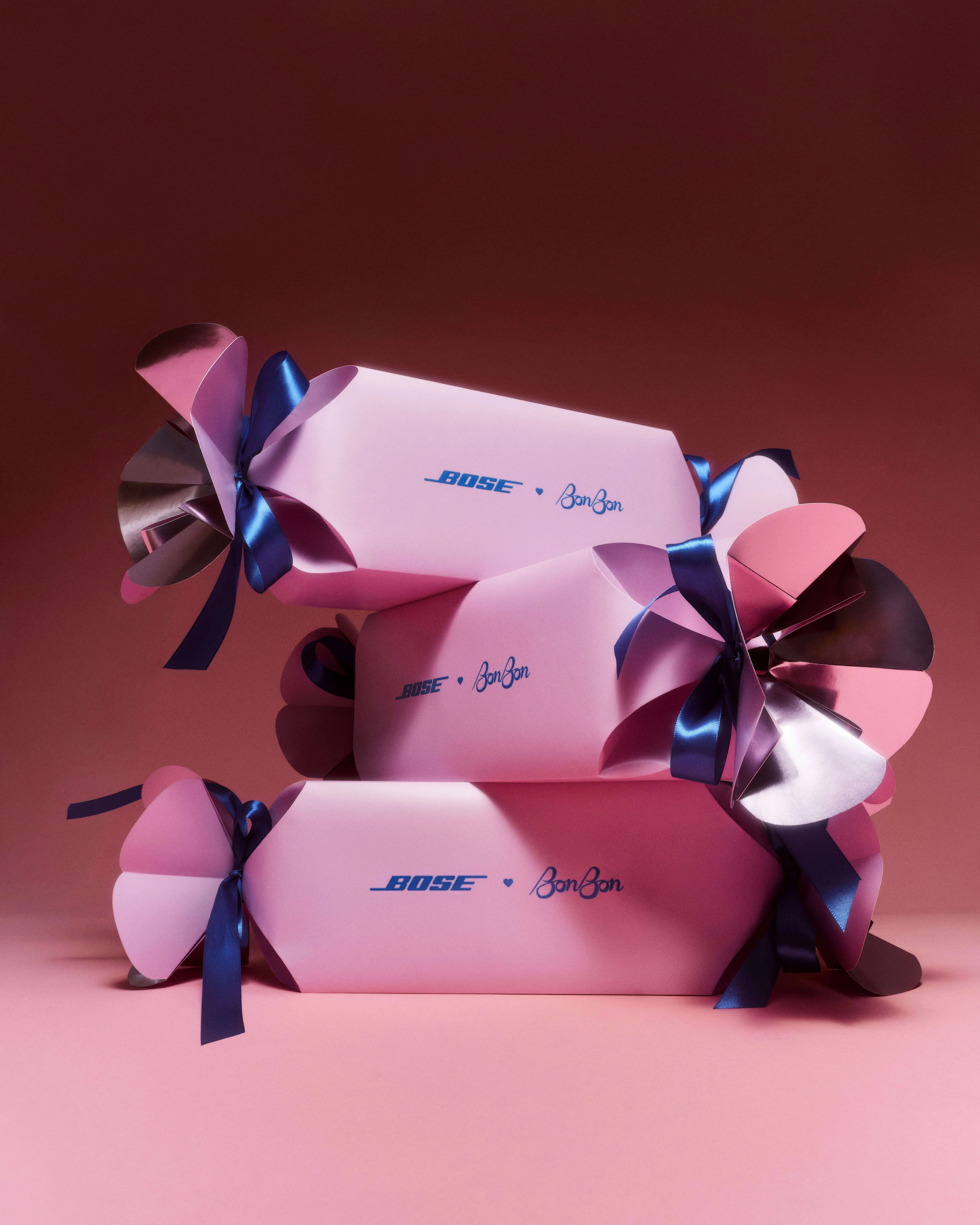 Three Bose × BonBon candy wrapper mailers stacked at varying angles, showing the full sculptural form with fanned paper ends and navy ribbon ties against a deep rose backdrop.