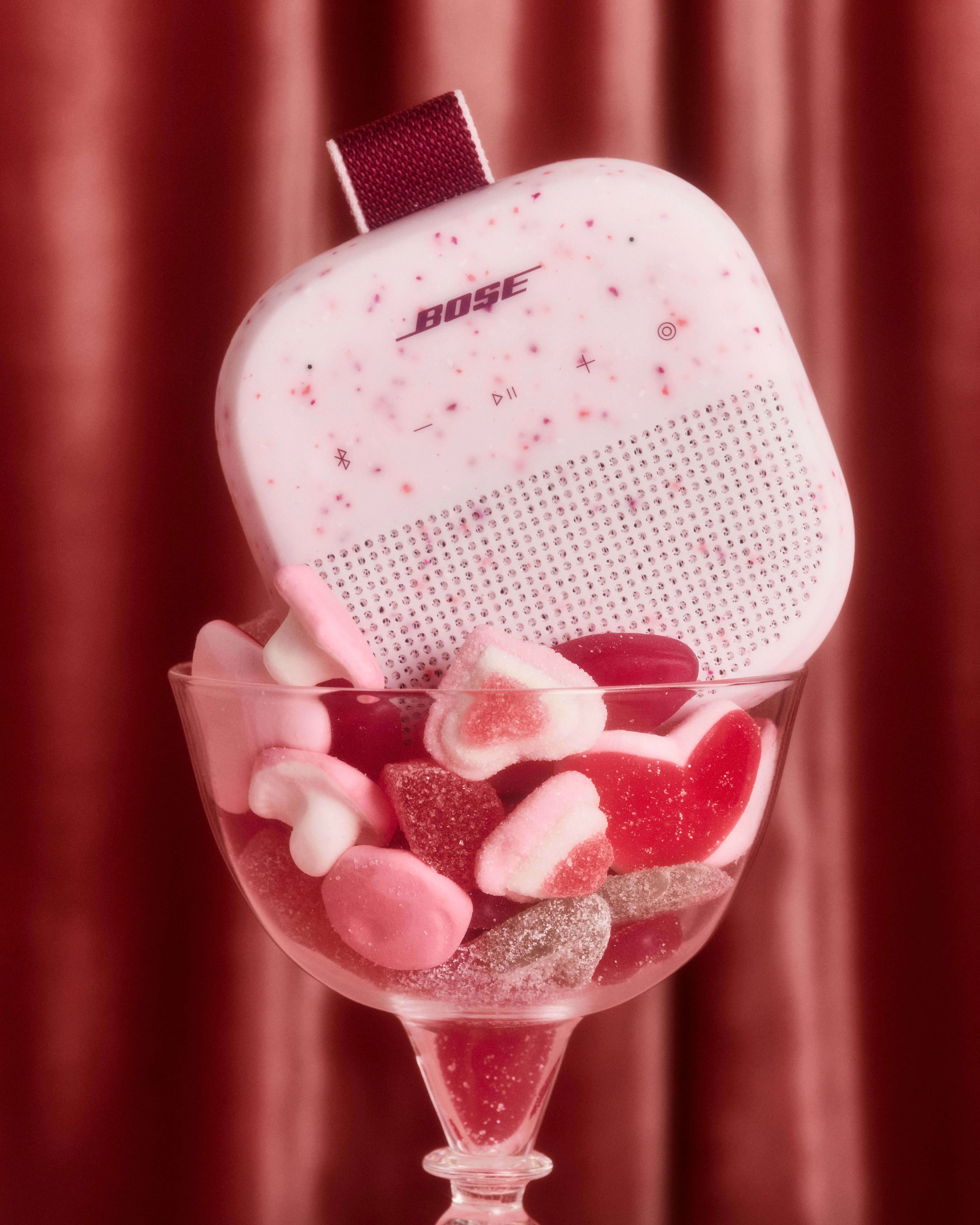 Bose Soundlink Micro in Petal Pink perched above a crystal coupe glass filled with Valentine's gummy candies, set against a deep red velvet curtain.