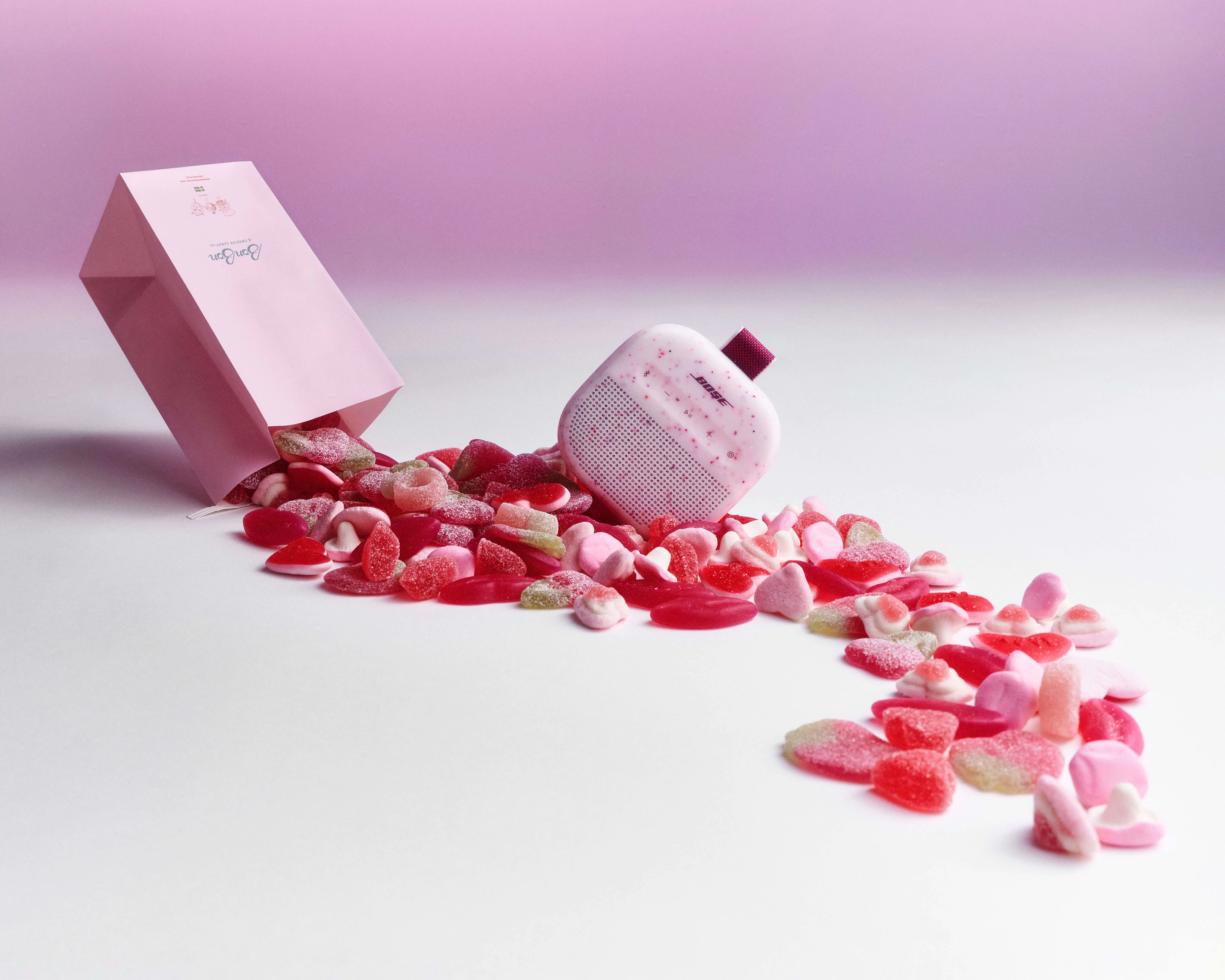 Bose Soundlink Micro in Petal Pink tipped on its side amid a scattered pile of Valentine's gummy candies spilling from an open pink BonBon bag.