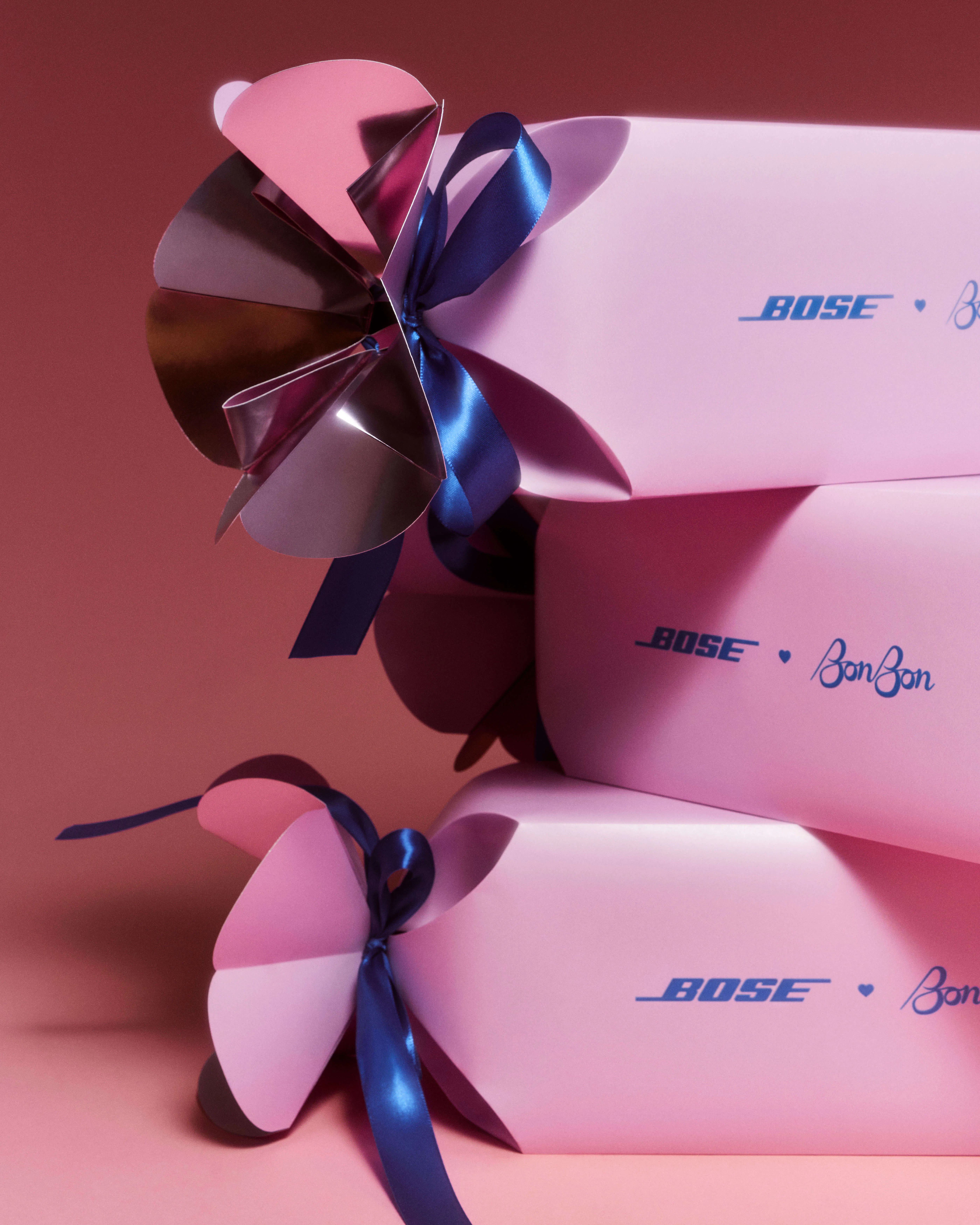 Close-up of three stacked Bose × BonBon candy wrapper mailers in pink, with engineered petal folds at each end tied with deep navy satin ribbon.
