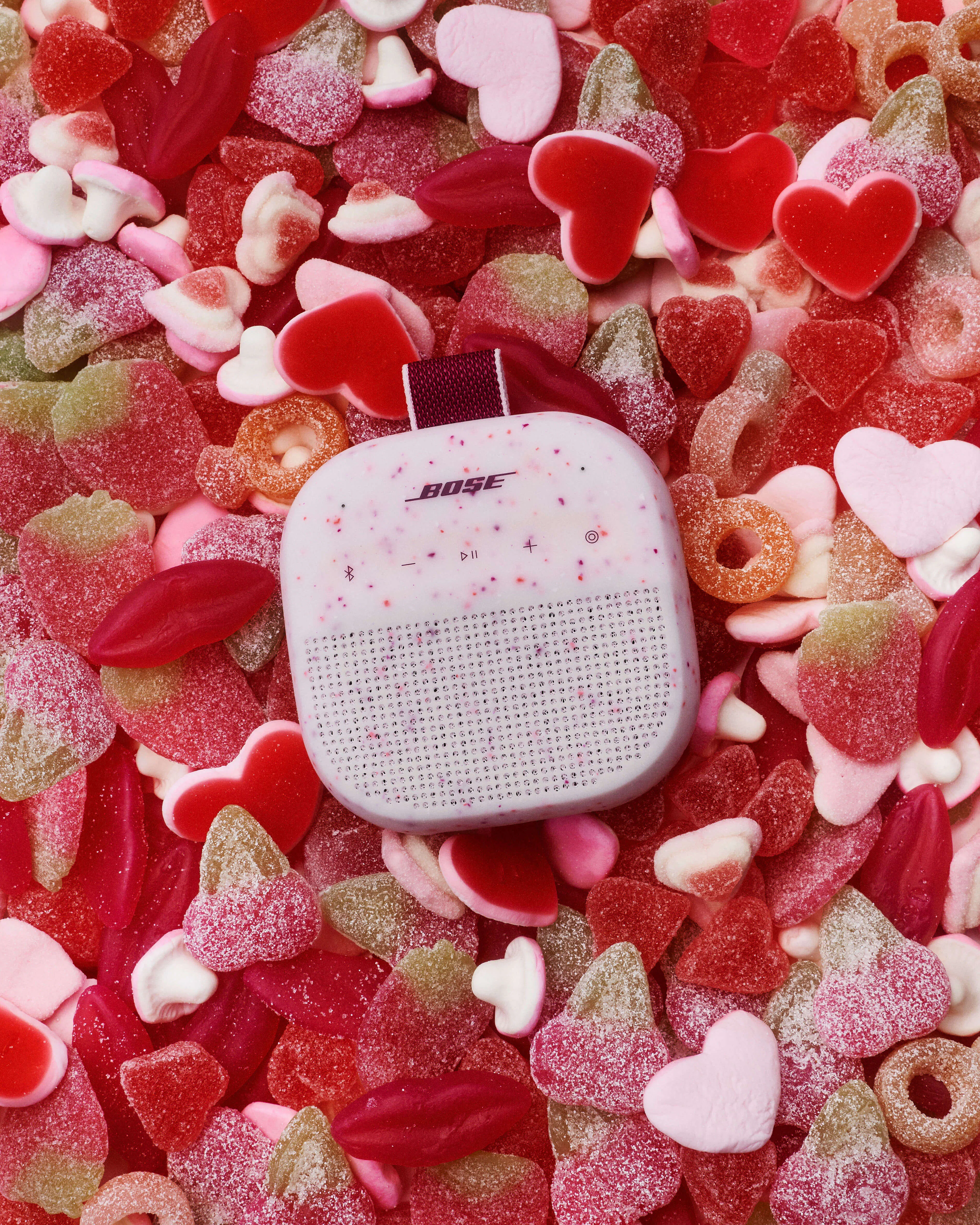 Overhead flatlay of the Bose Soundlink Micro in Petal Pink surrounded by a dense spread of Valentine's gummy candies in red, pink, and white heart shapes.