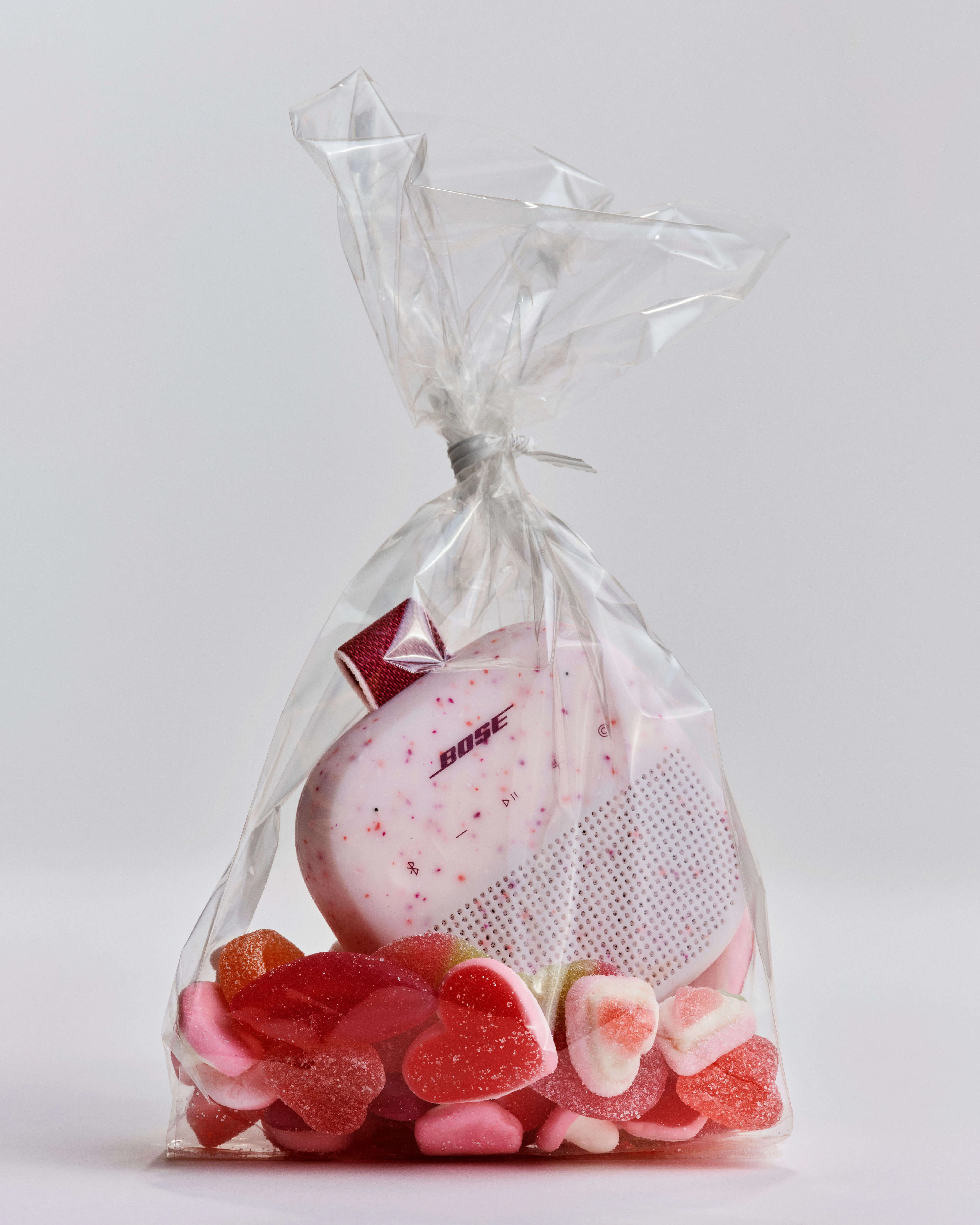 Bose Soundlink Micro in Petal Pink nestled among heart-shaped and assorted Valentine's gummy candies inside a clear cellophane bag, tied at the top.