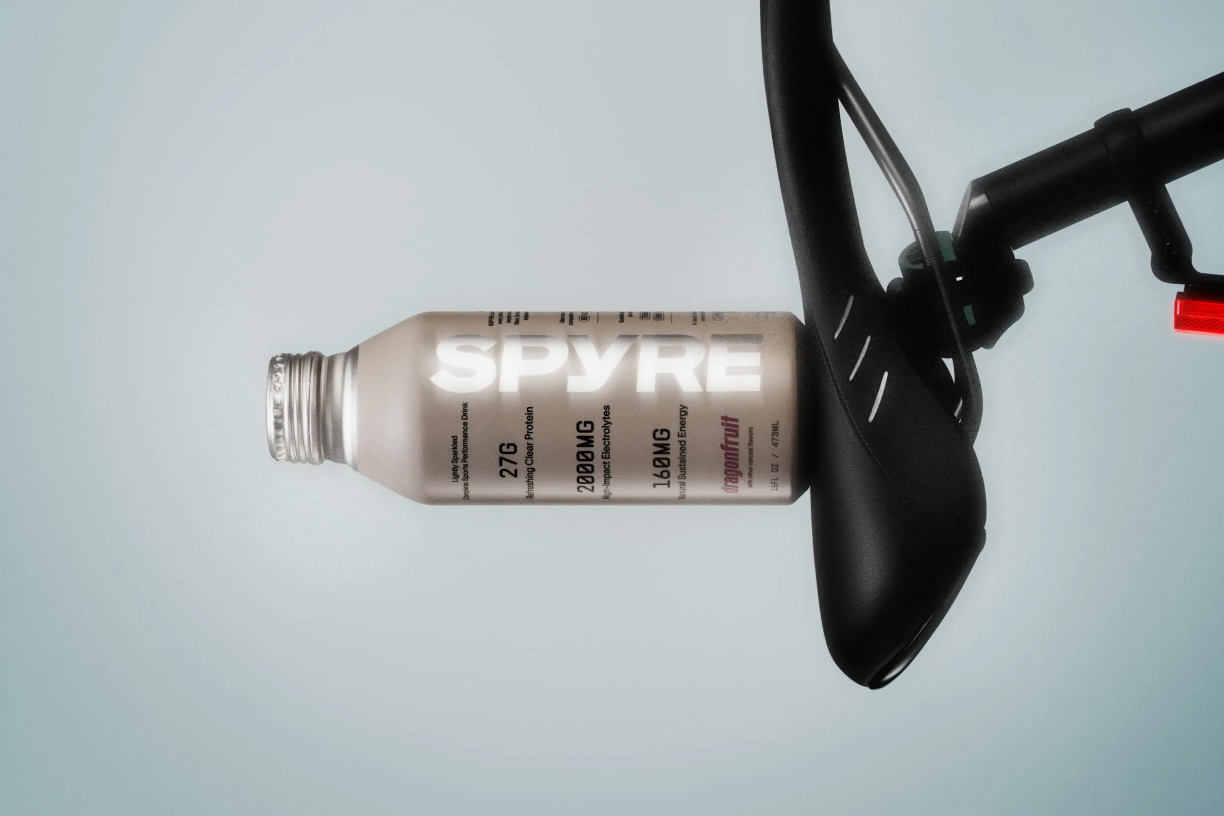 Overhead flat-lay of a SPYRE dragonfruit performance drink bottle lying horizontally next to a sleek black road bike saddle and seatpost, against a pale grey-blue background