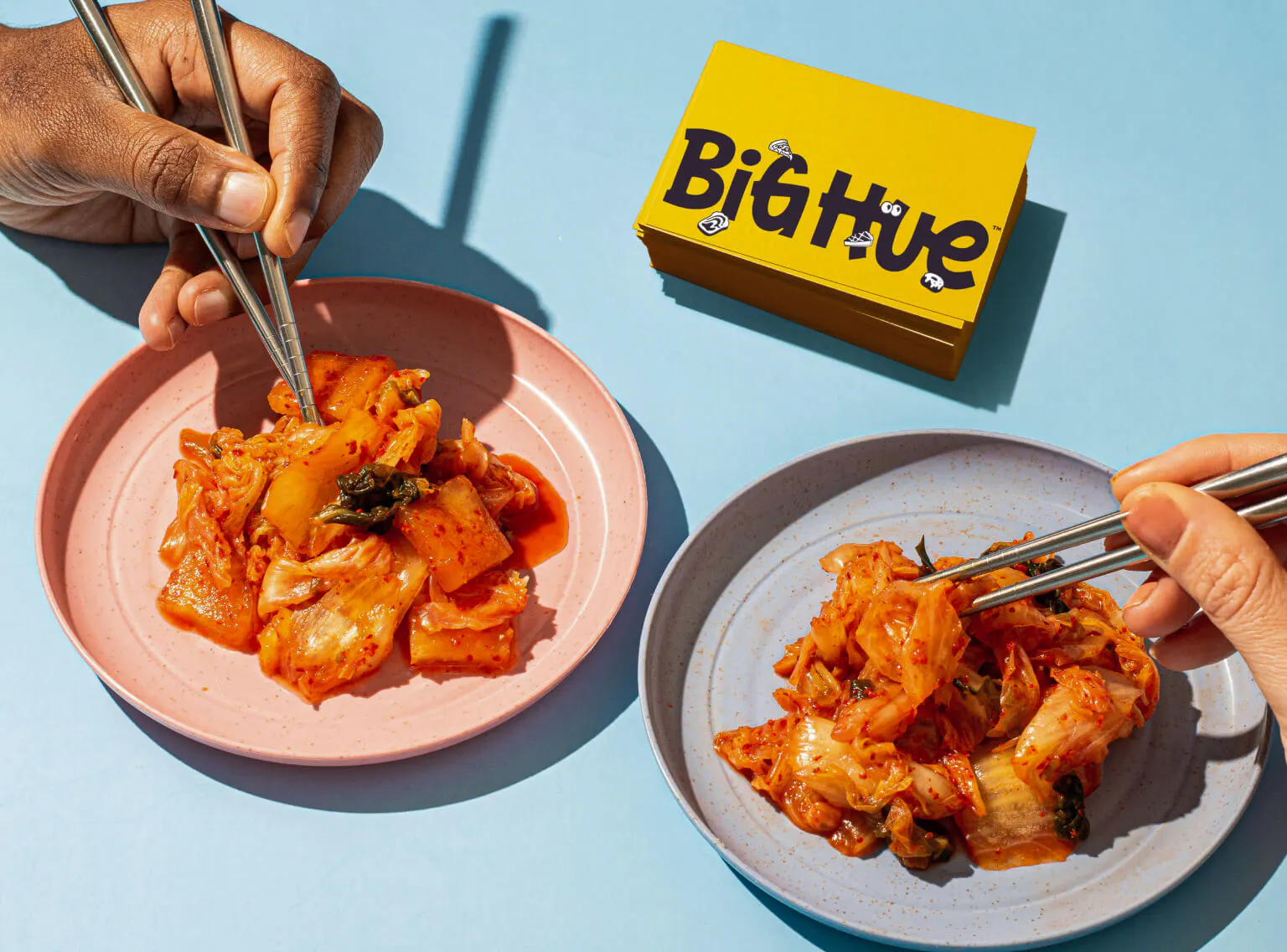 Two hands holding chopsticks over plates of kimchi on a light blue surface. A stack of yellow Big Hue business cards sits in the upper right corner.