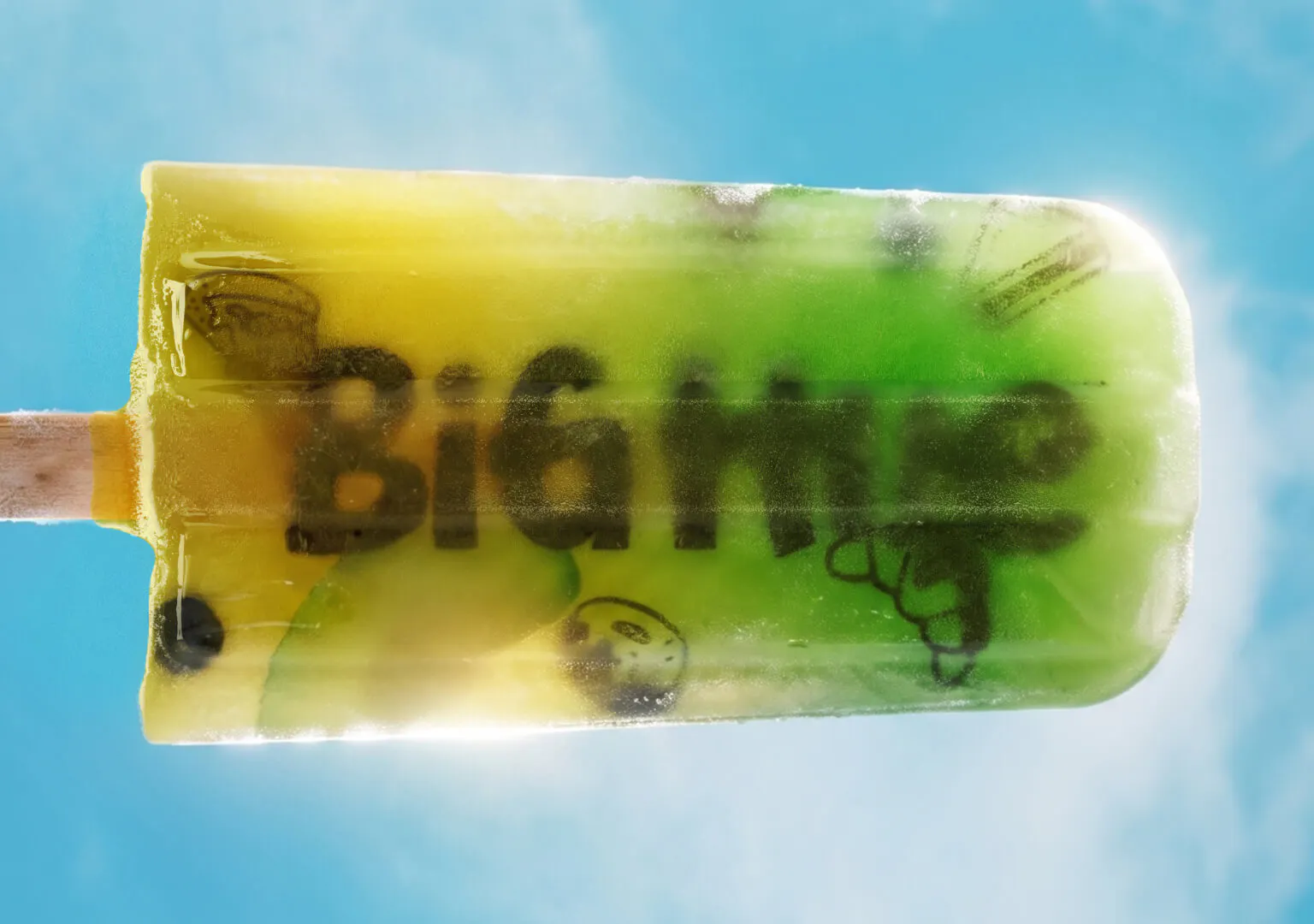 A yellow-to-green gradient popsicle held against a bright blue sky. The Big Hue wordmark and food illustrations are visible frozen inside the ice.