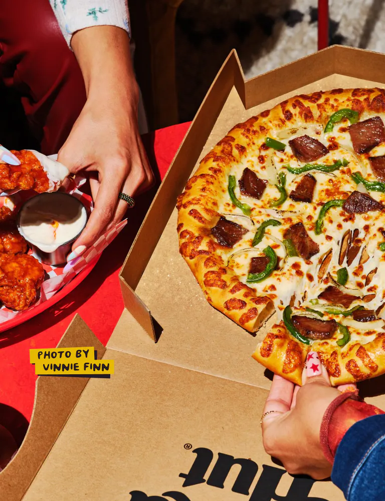 Overhead shot of two hands — one holding a tray of boneless wings, one pulling a cheesy pizza slice from an open Pizza Hut box. A yellow label reads "Photo by Vinnie Finn."